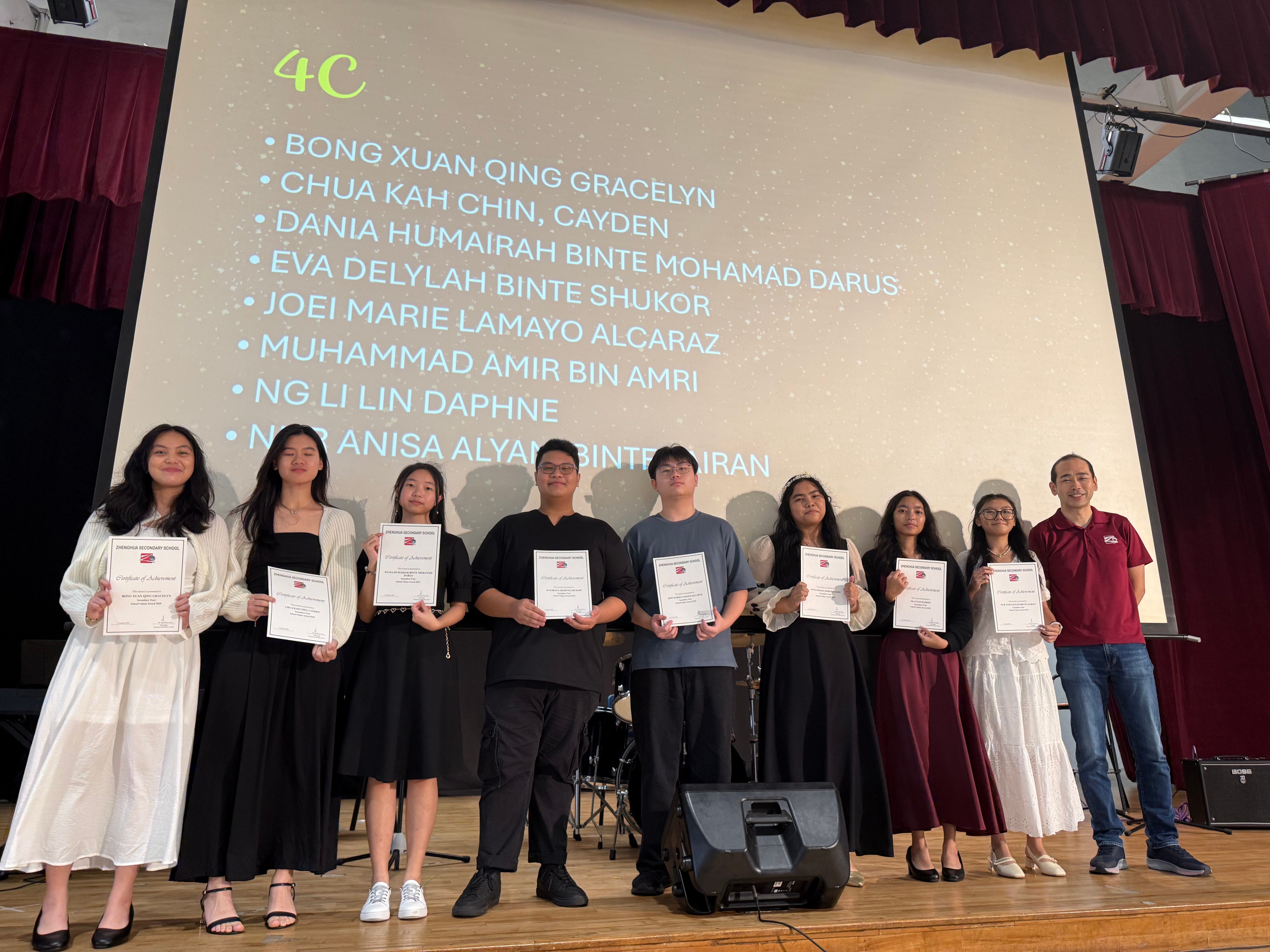 Nine students and a teacher stand on a stage holding certificates, with a class 4C names list projected behind them.