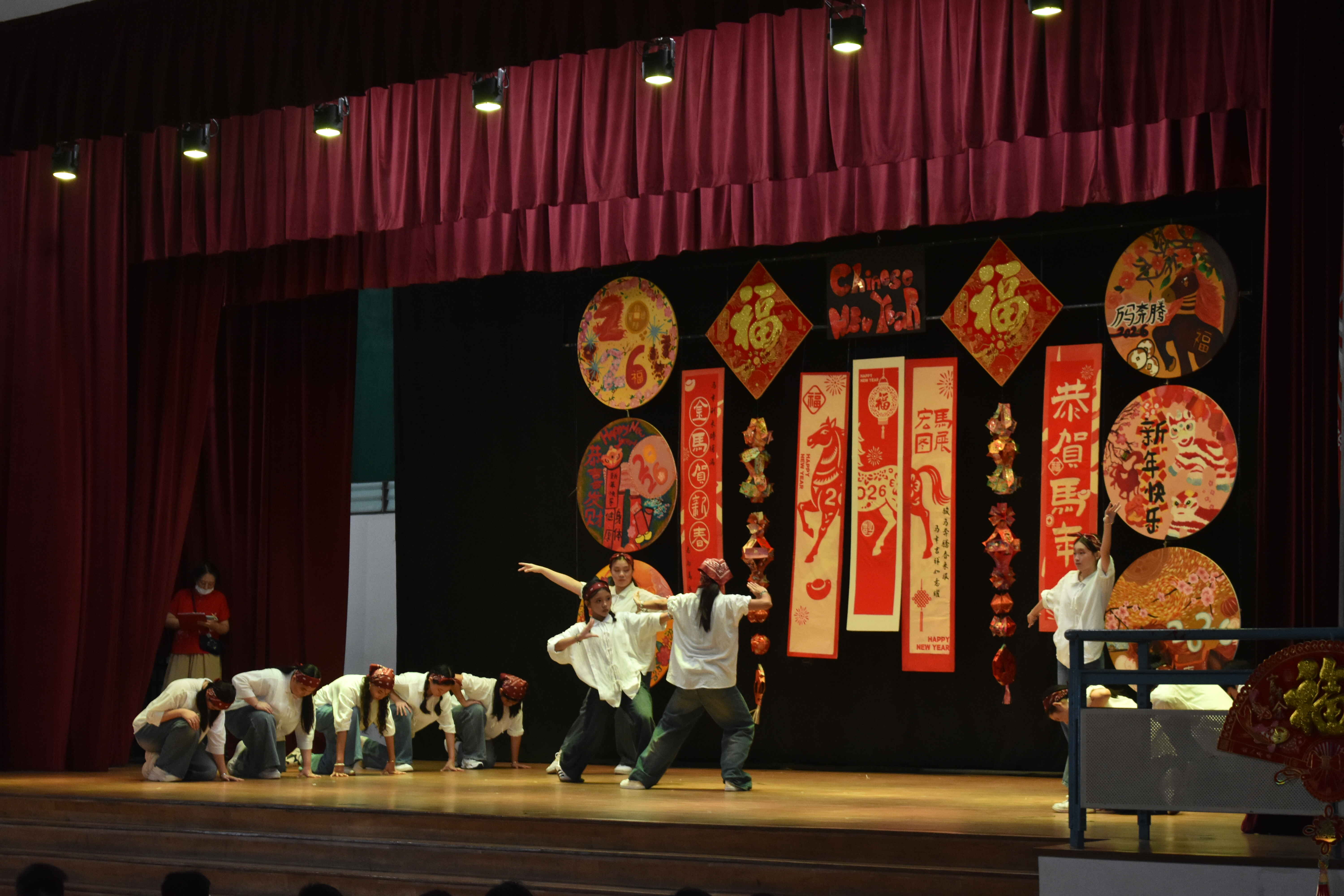 Students in white shirts perform a dance on a stage decorated with red Chinese New Year banners and circular artworks.