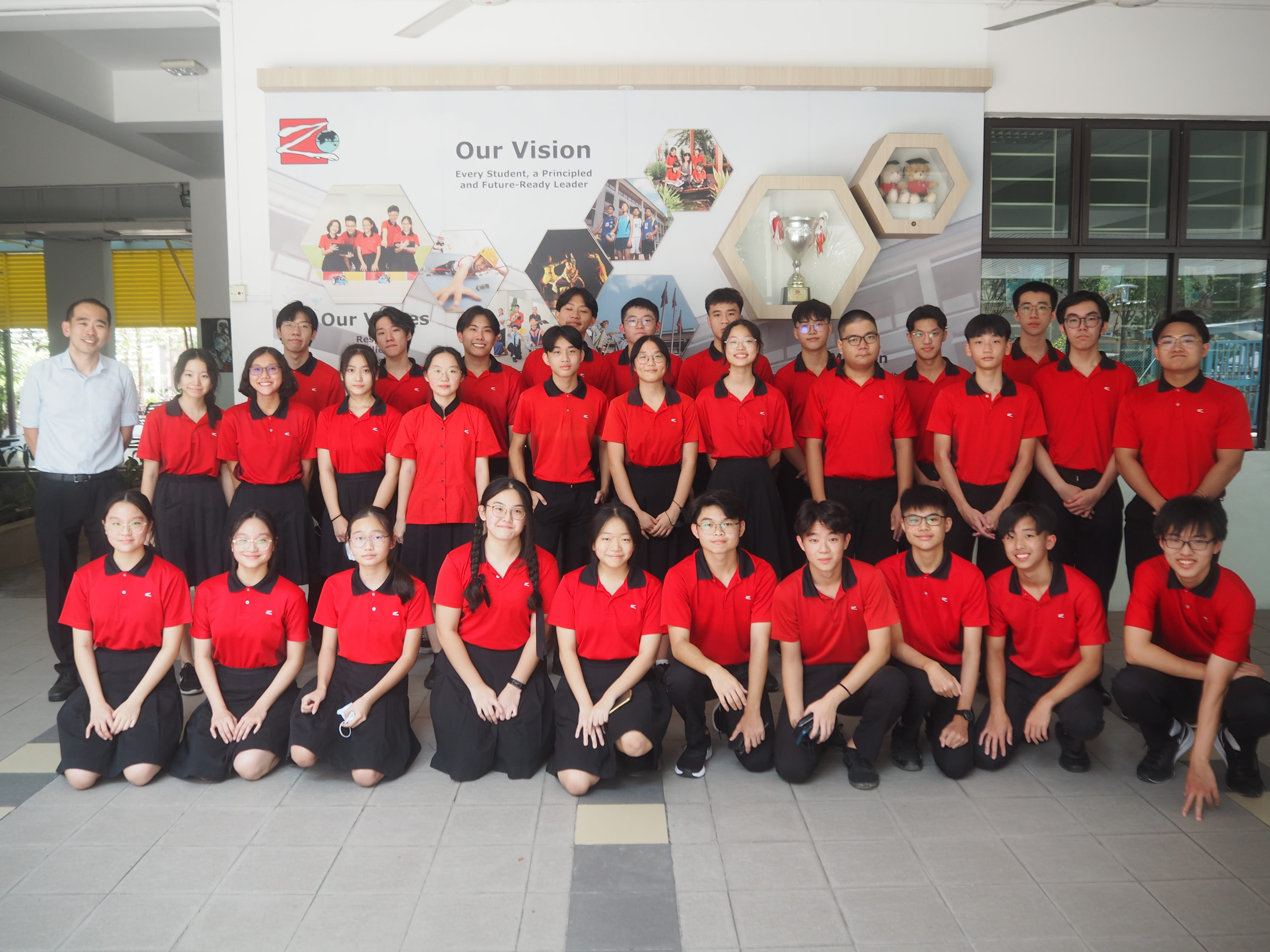 A large group of students in red and black uniforms pose in three rows before a school vision display board.