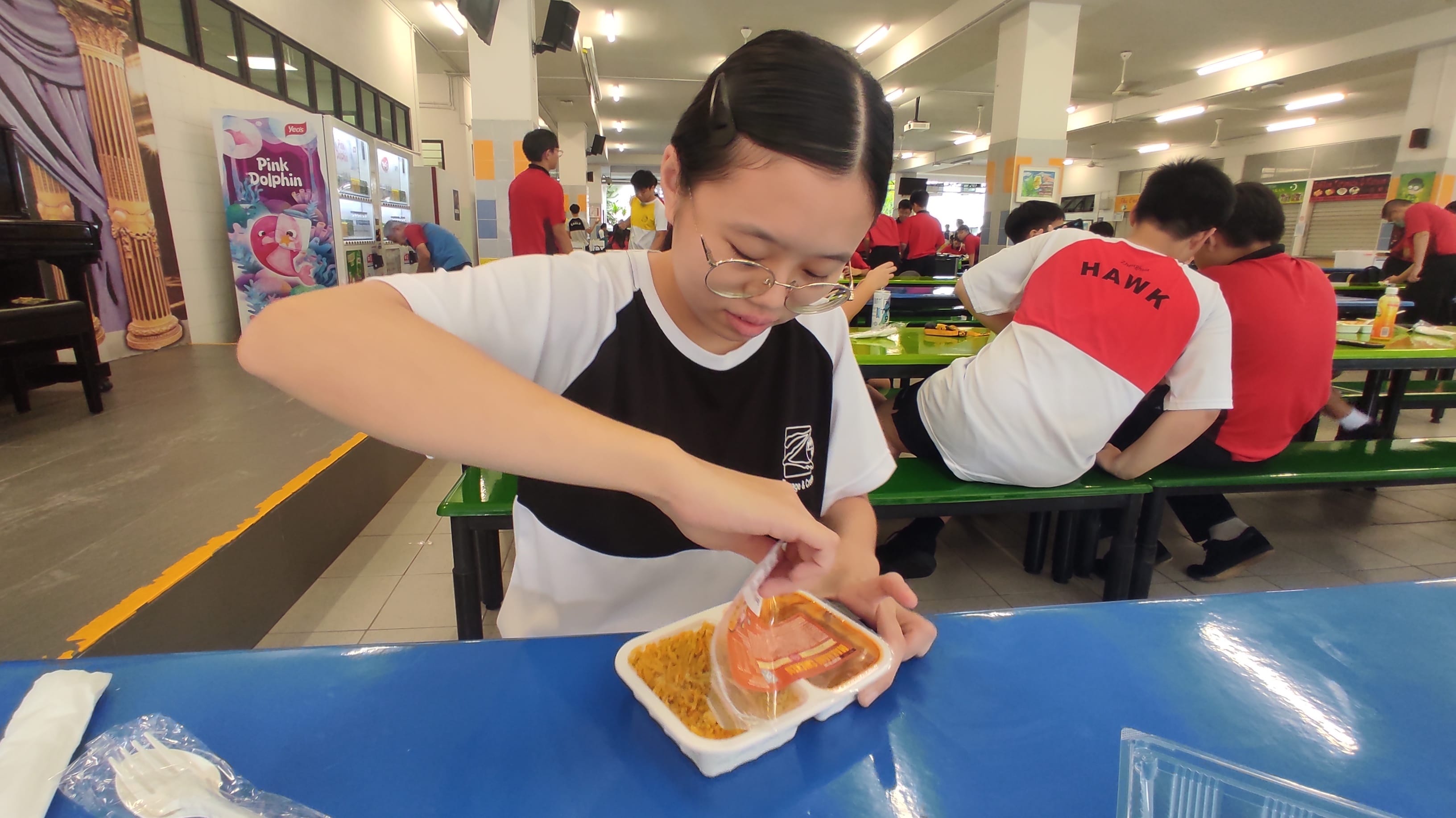 A girl with glasses opens a plastic food container at a school canteen table with students eating in the background.