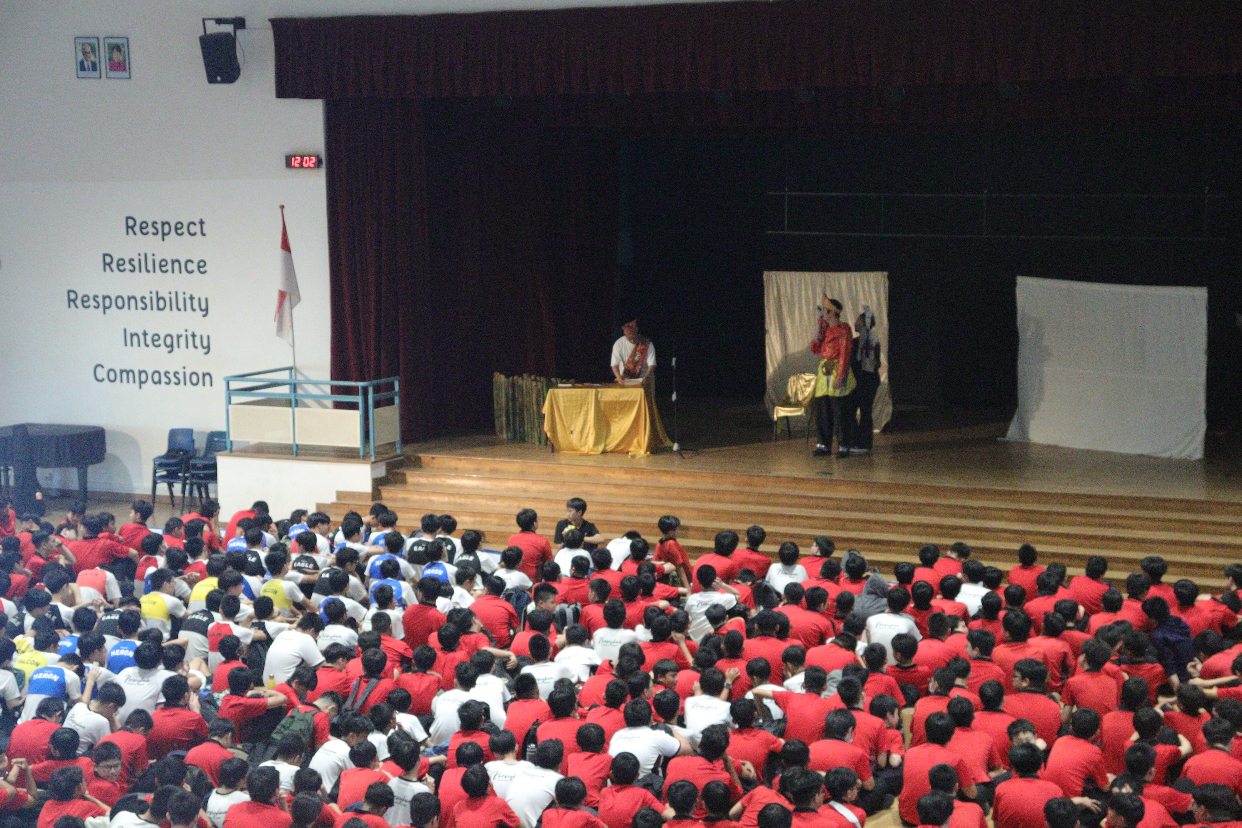 Large school assembly hall with students in red and white shirts seated on floor watching two performers on a wooden stage.