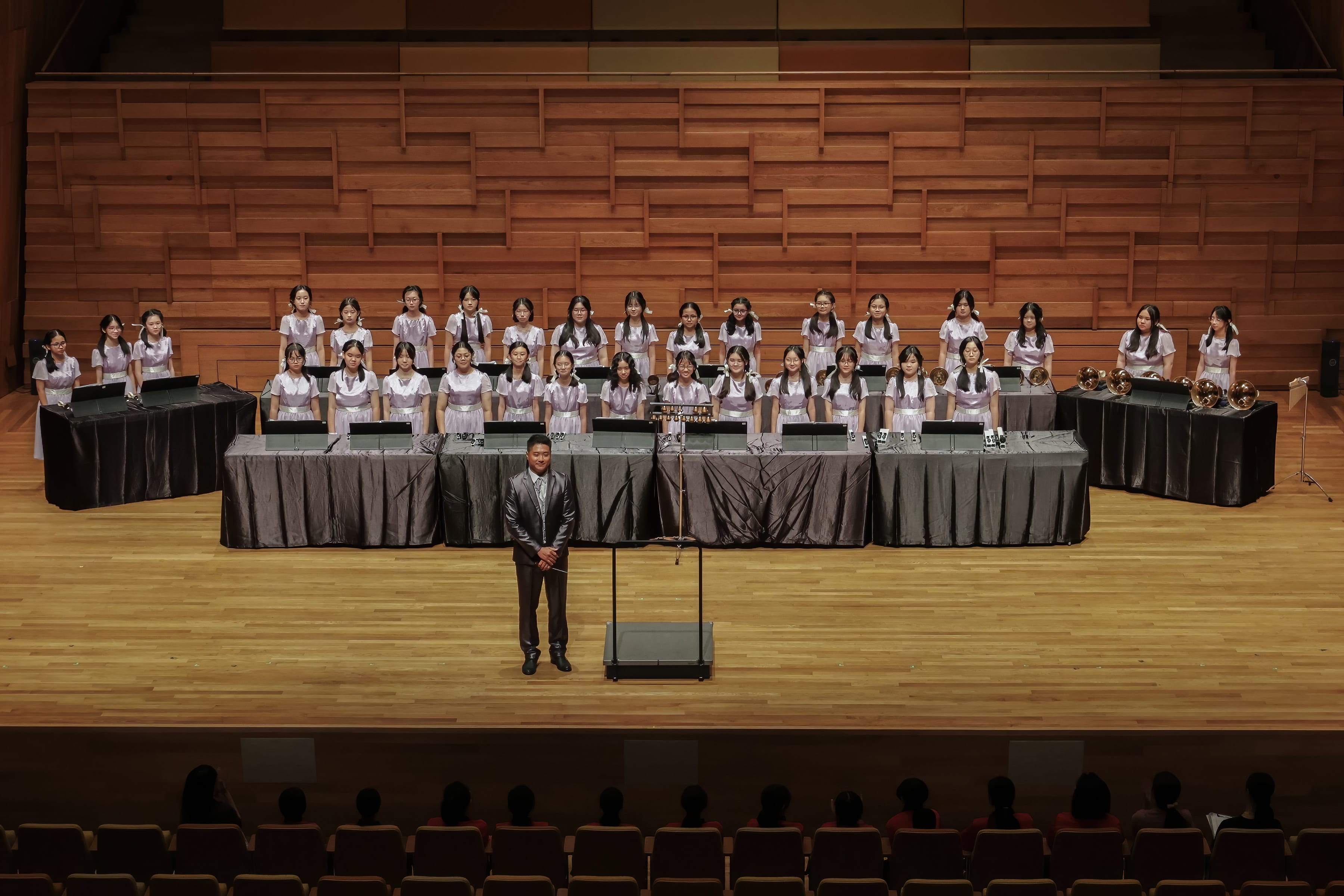A conductor in a suit stands before rows of young female musicians in matching dresses on a wooden concert stage.