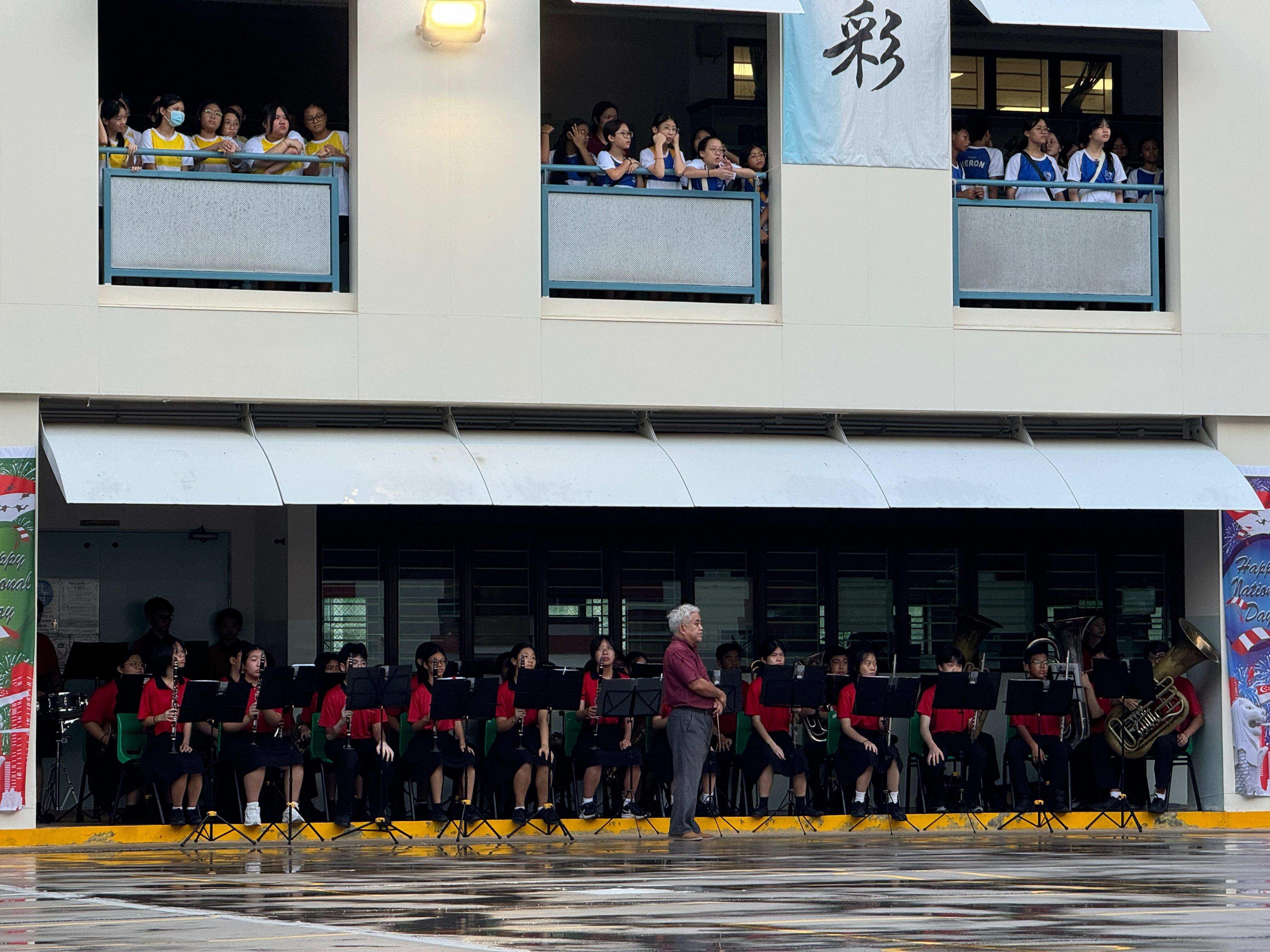 A conductor leads a school band in red uniforms outdoors while students watch from upper floor balconies above.