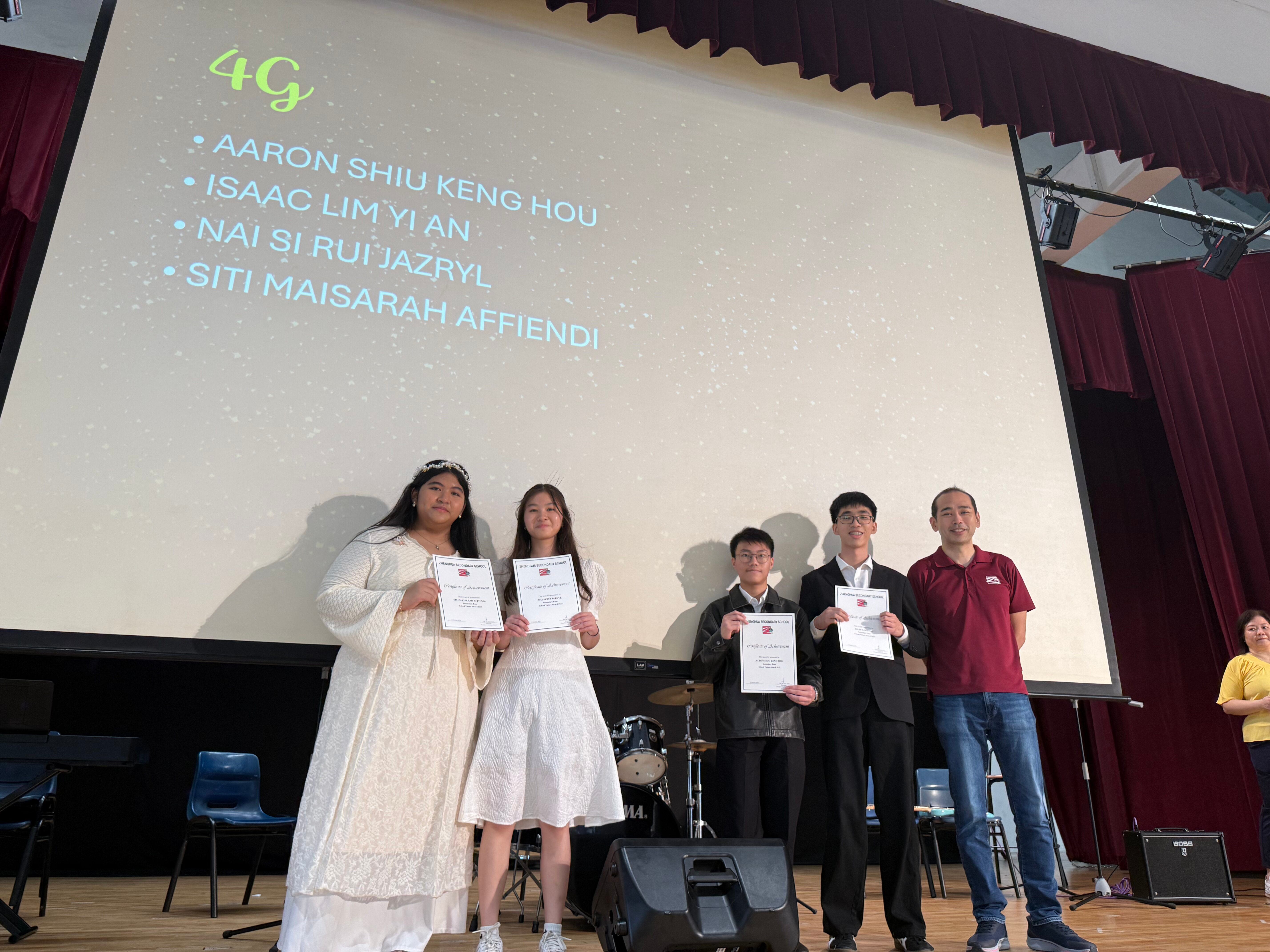 Four students holding certificates stand on stage with a teacher, a screen behind them lists class 4G student names.