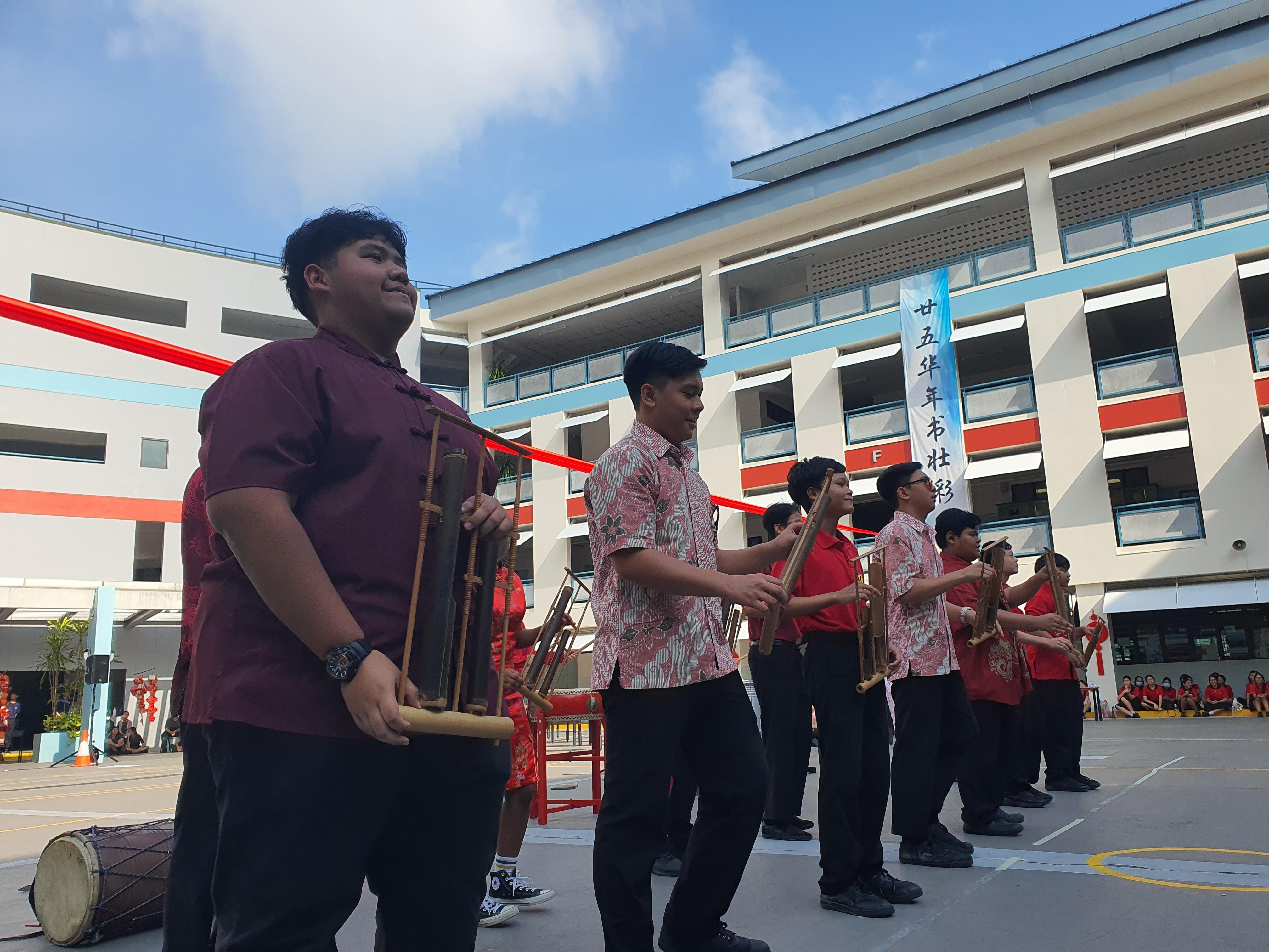 Students in red and maroon shirts play bamboo instruments in a school courtyard with Chinese banners visible.