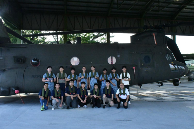 A group of young people in military uniforms pose in front of a large dark green tandem-rotor helicopter in a hangar.