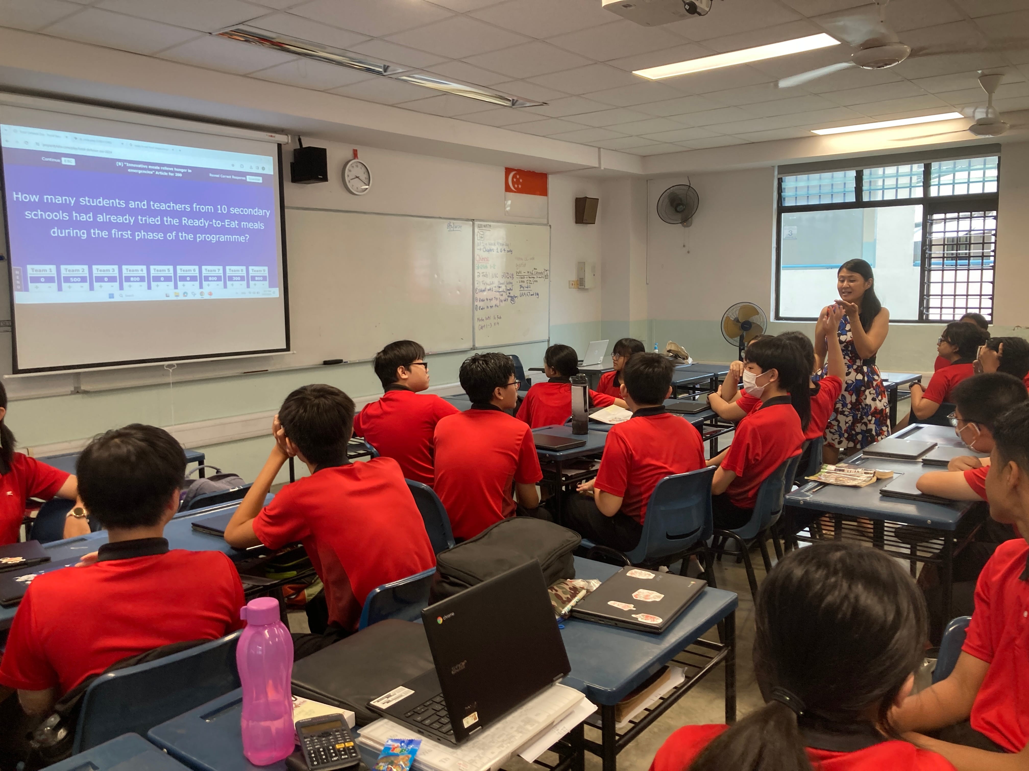 A teacher stands beside a projected quiz question while students in red uniforms sit at desks with laptops.
