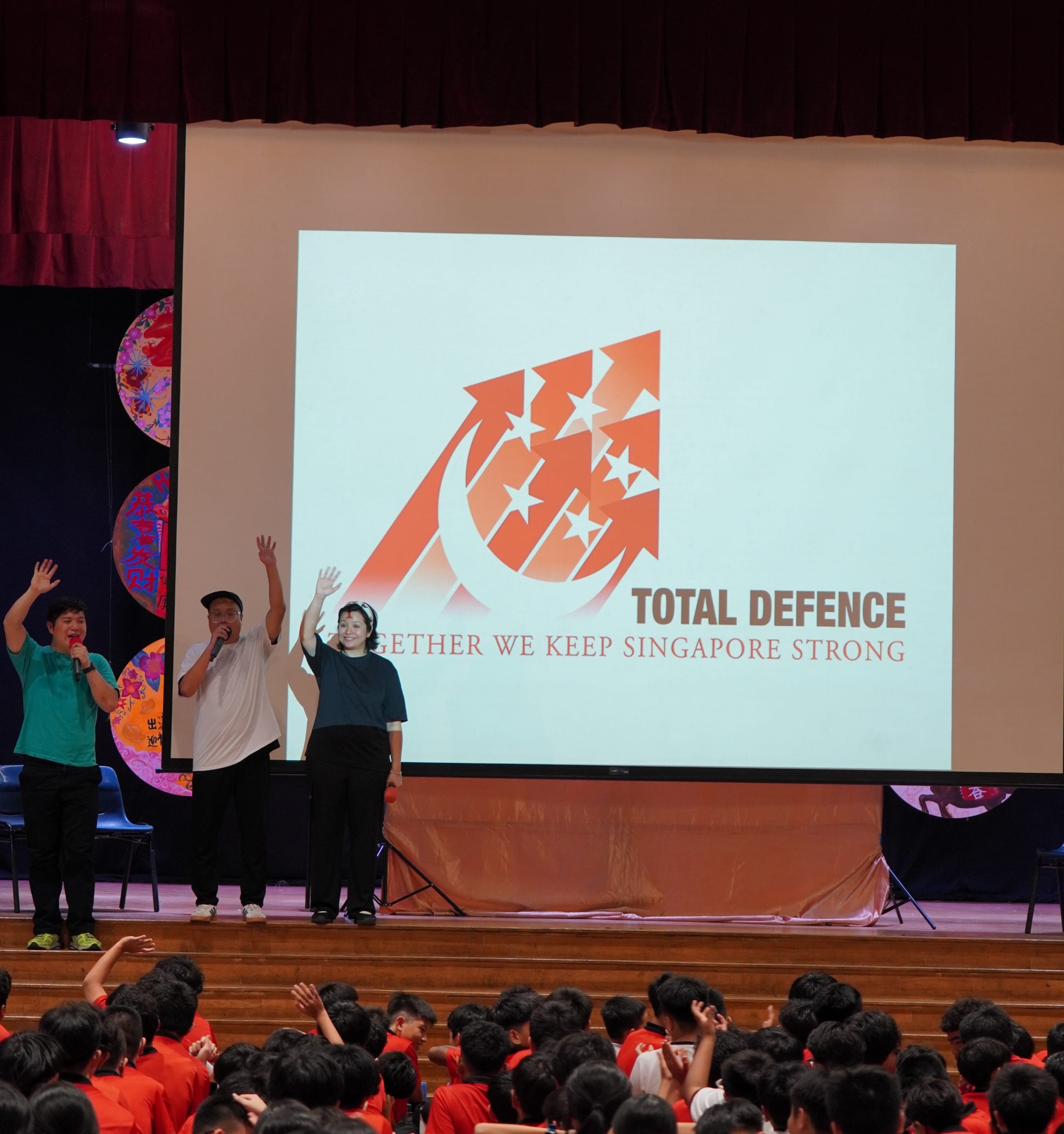 Three presenters on stage wave to a student audience before a Total Defence Singapore projection.