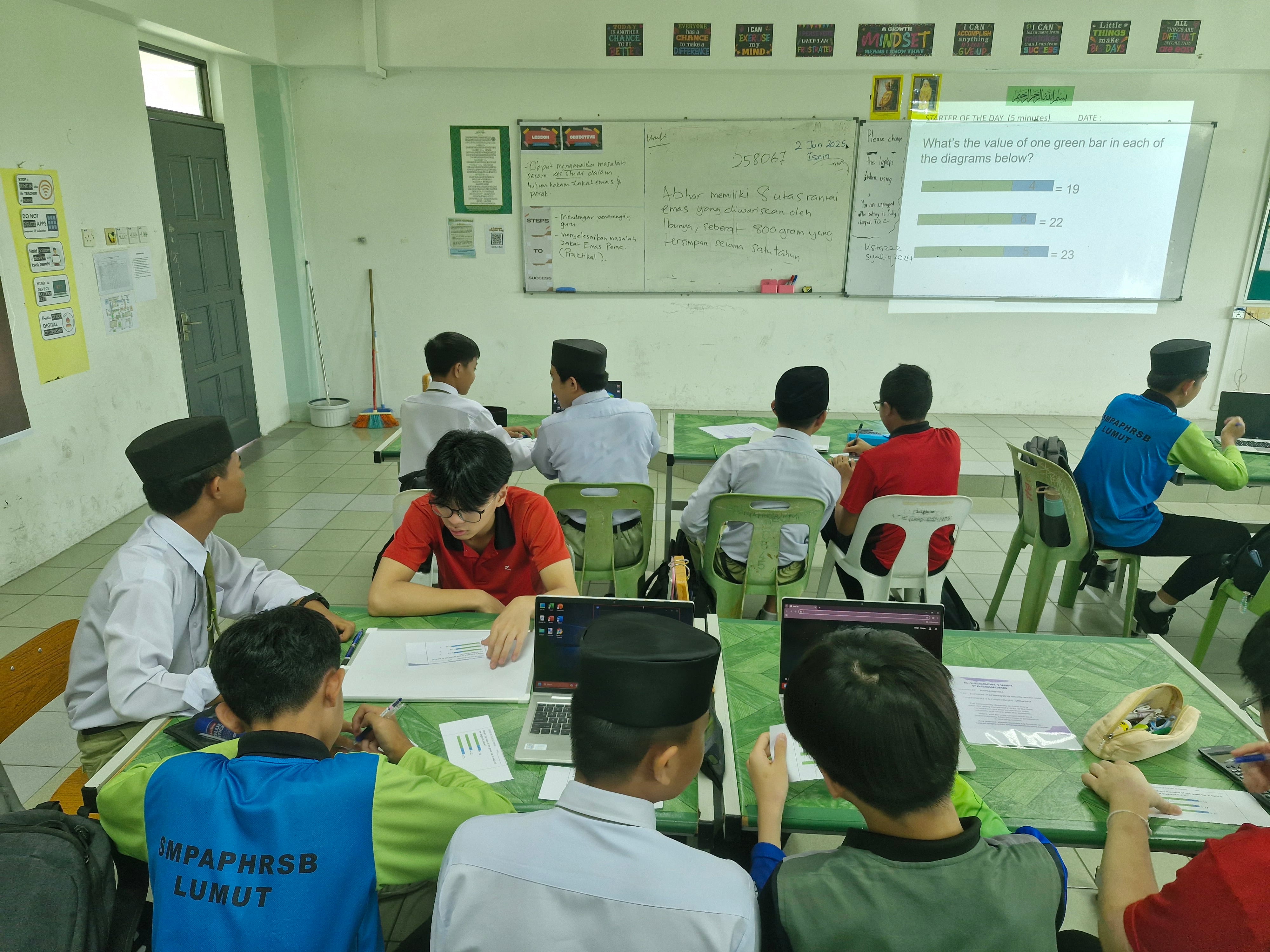 Students sit at tables in a classroom, working on math while a projector shows bar diagrams on the board.