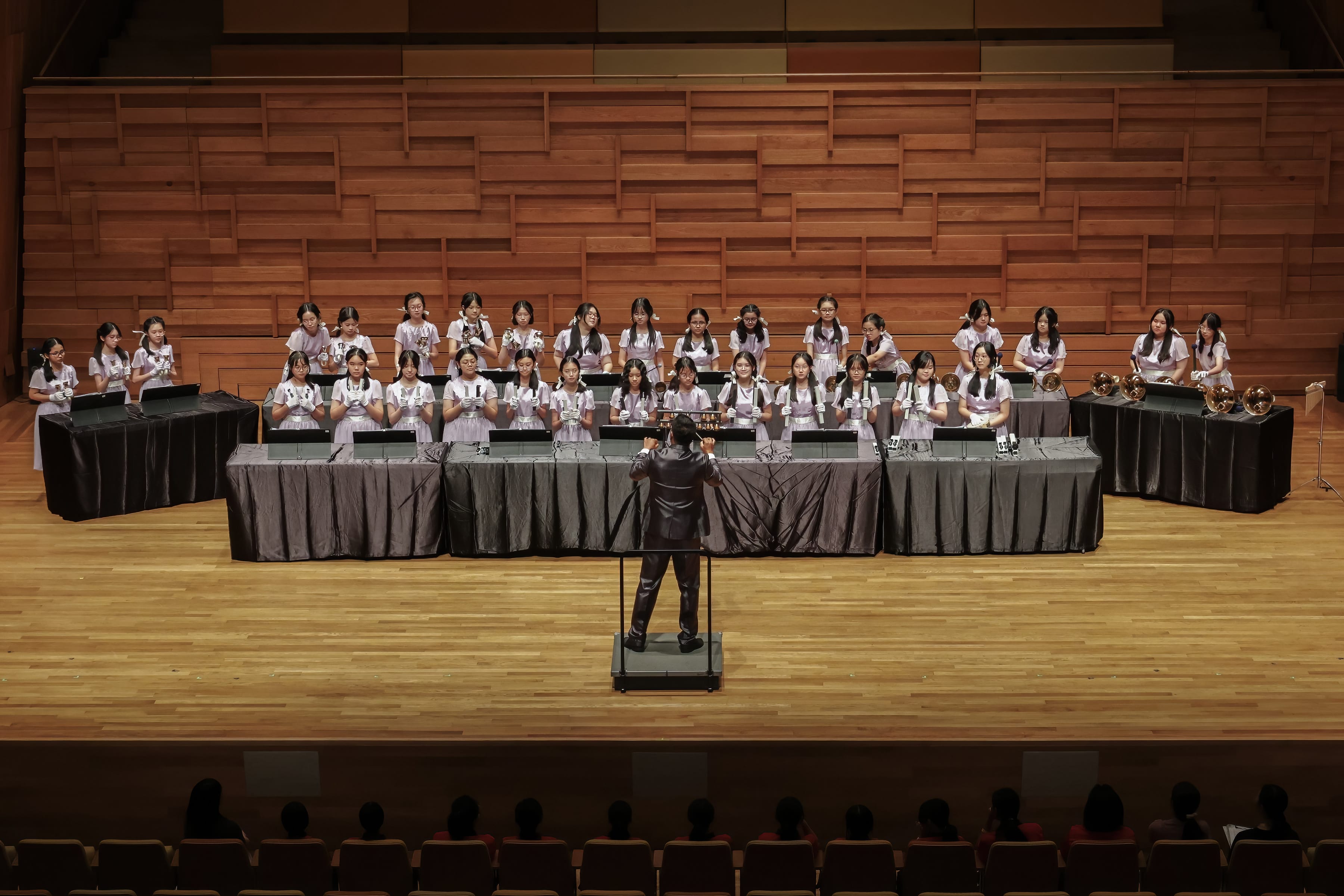 A conductor leads rows of young female musicians in white uniforms performing on a wooden concert hall stage.