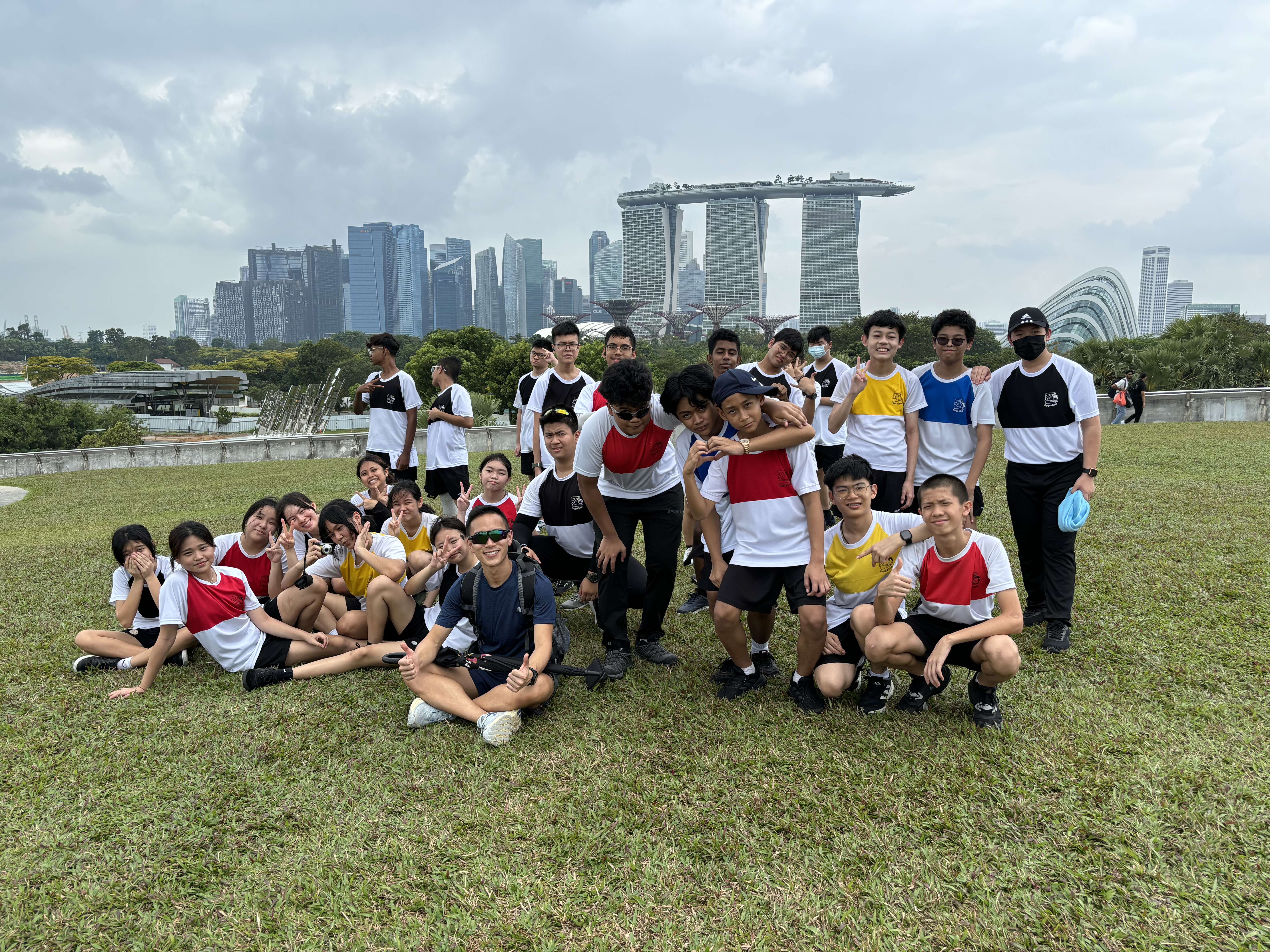 A group of about 20 students posing on a grassy hill with the Singapore skyline and Marina Bay Sands behind them.
