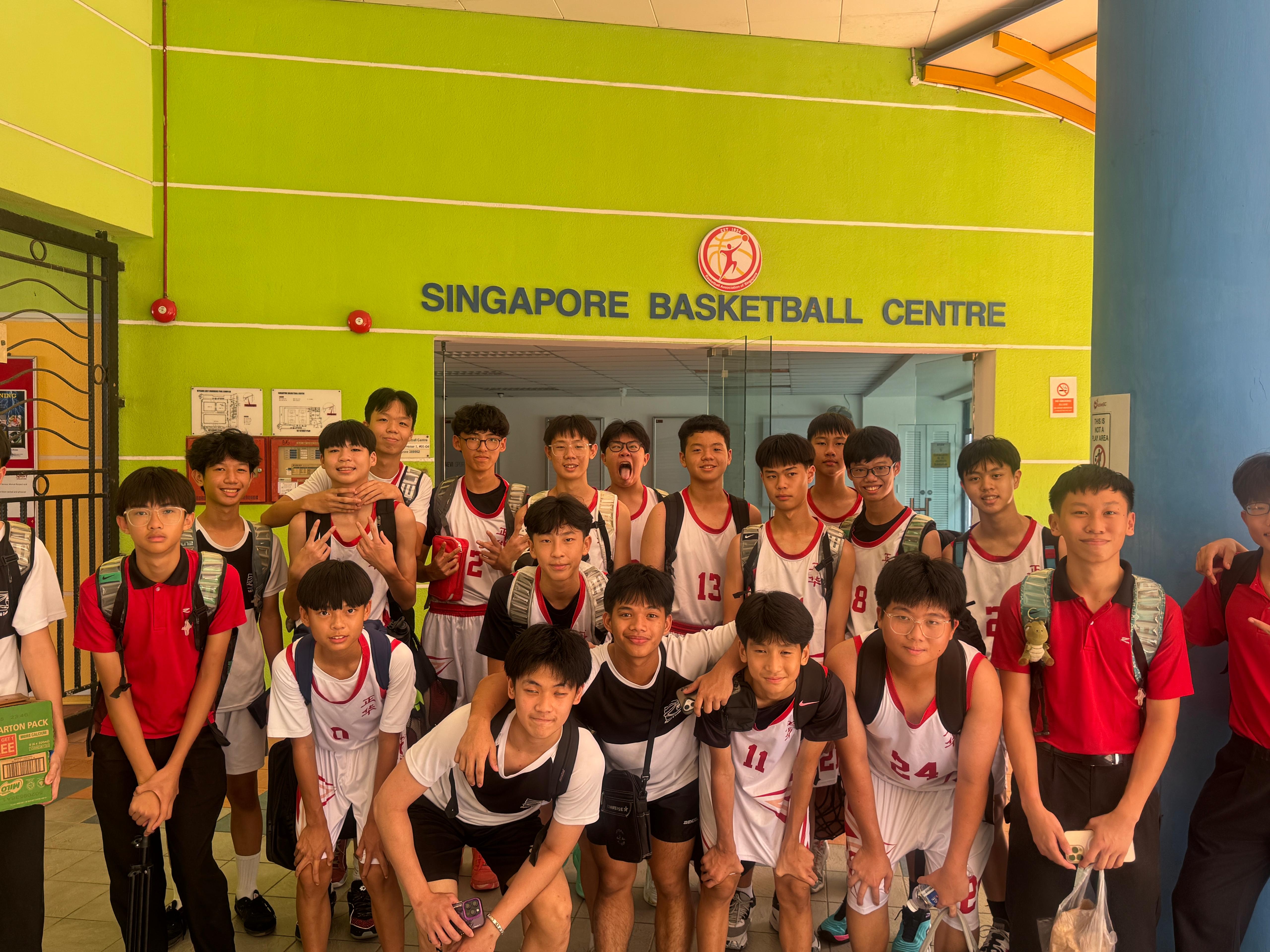 A group of teenage boys in basketball uniforms posing in front of the Singapore Basketball Centre entrance.