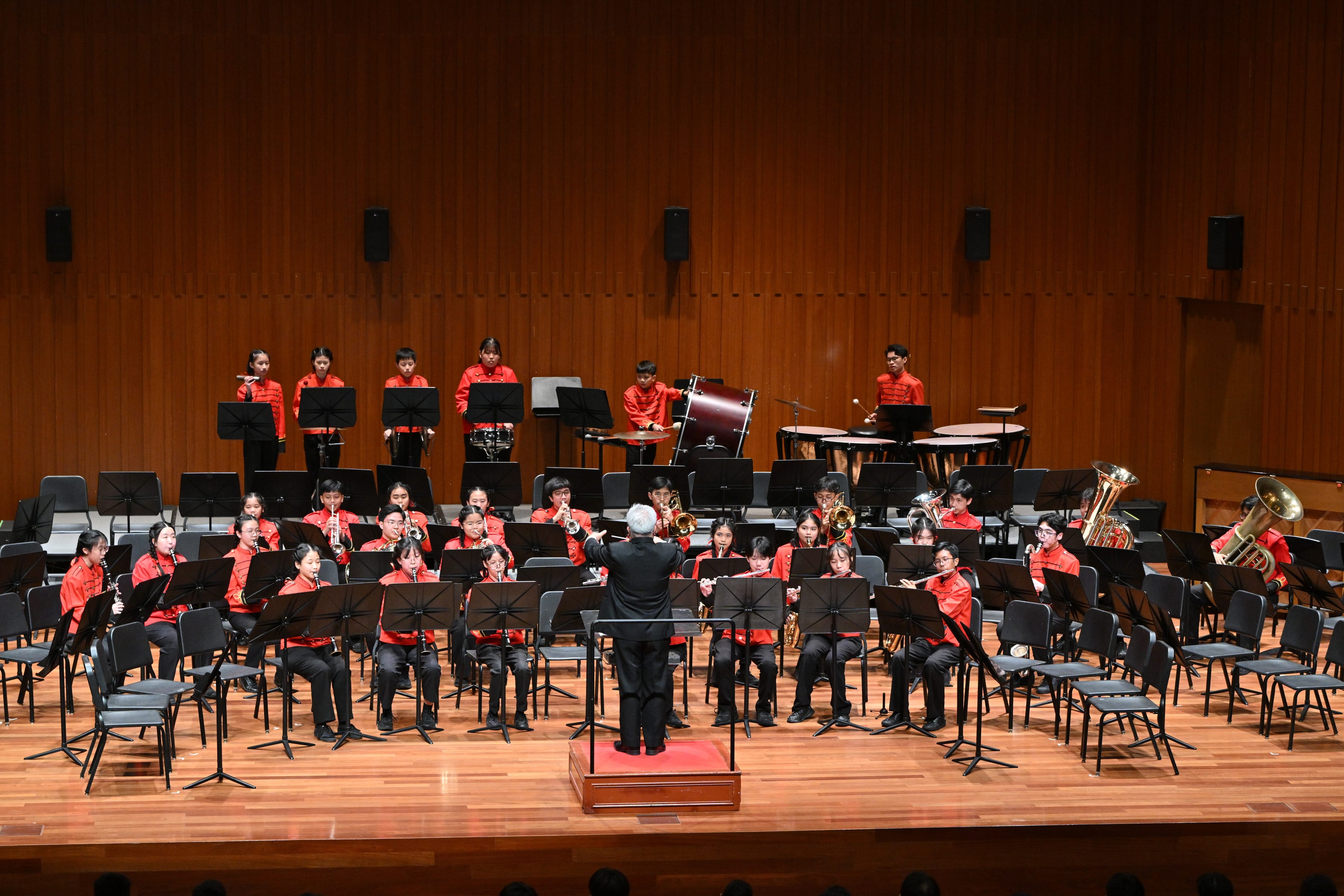 Conductor leads youth concert band in red shirts on a wooden stage with music stands and percussion in back.