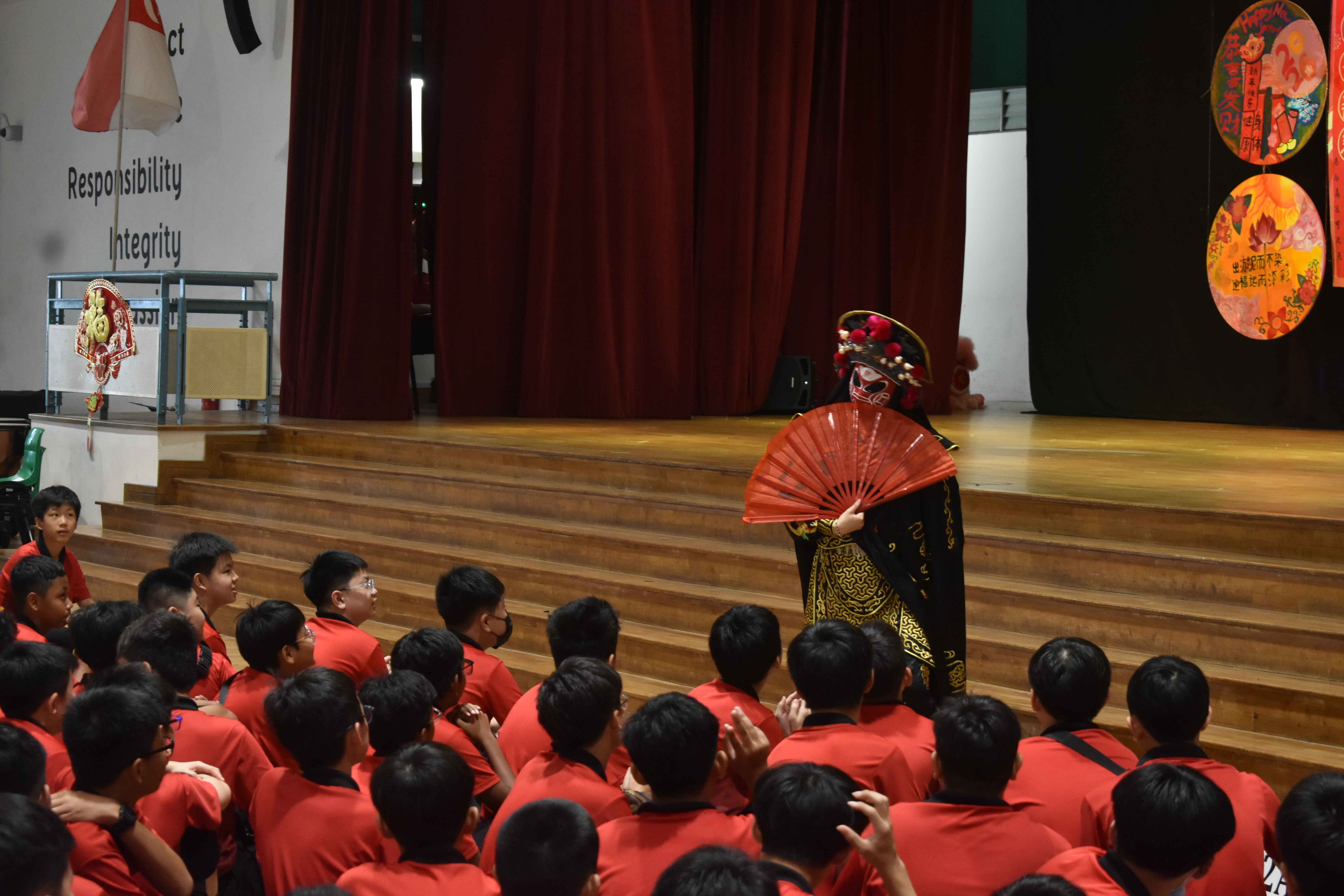 A masked performer in traditional Chinese opera costume holds a red fan before a seated crowd of children in red shirts.