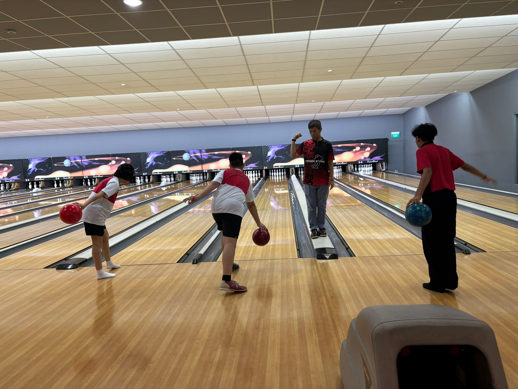 Four people at a bowling alley, three holding balls on wooden lanes with pins visible in the distance.