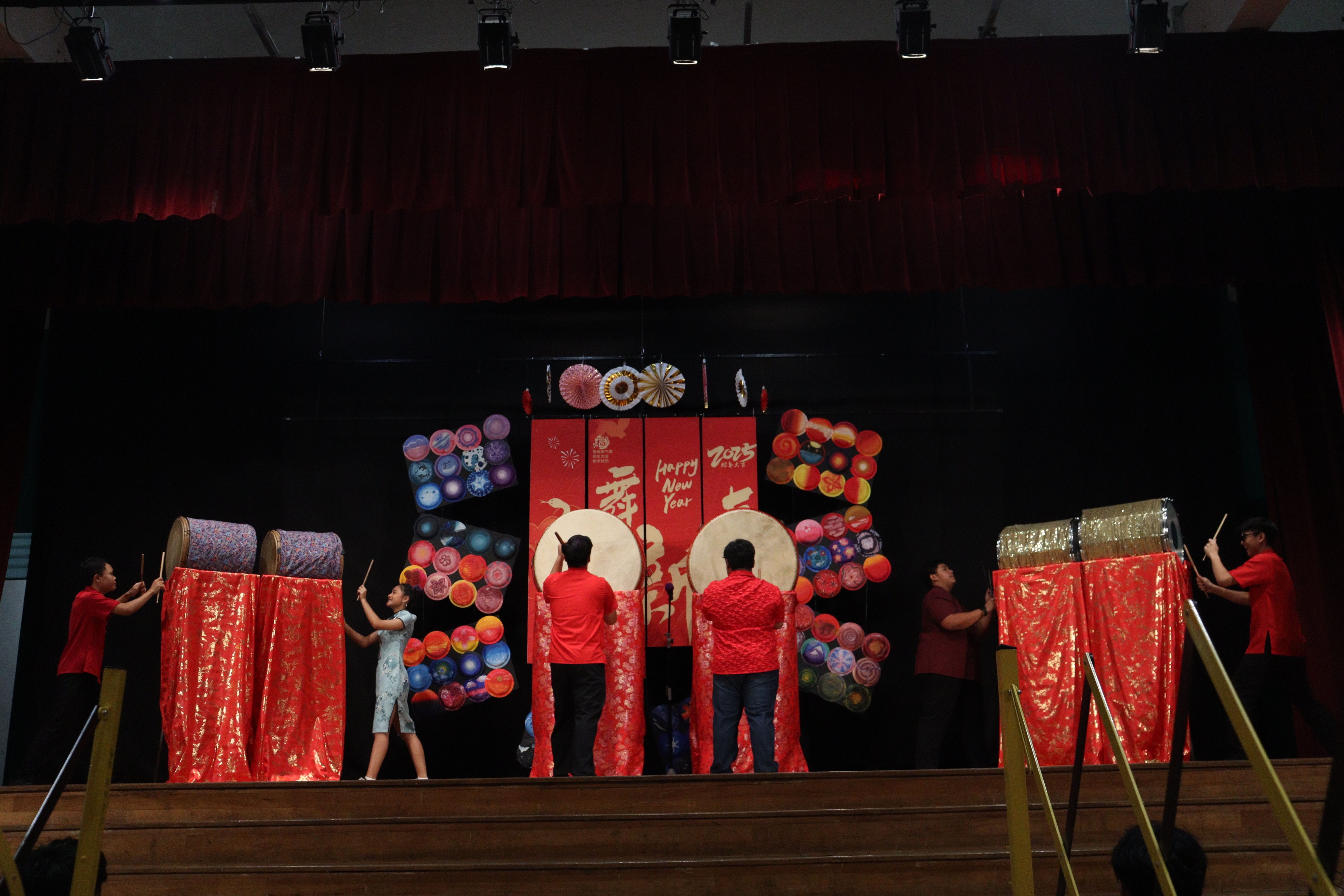 Five people in red outfits perform on a stage decorated with drums and a Happy New Year 2025 backdrop.