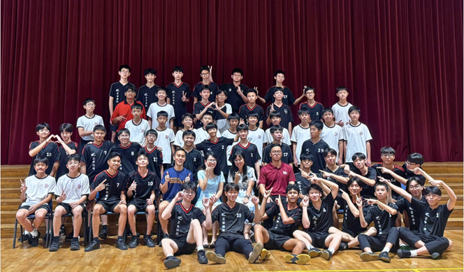 Large group of young students in black and white uniforms posing together on a wooden stage floor with a red curtain backdrop.