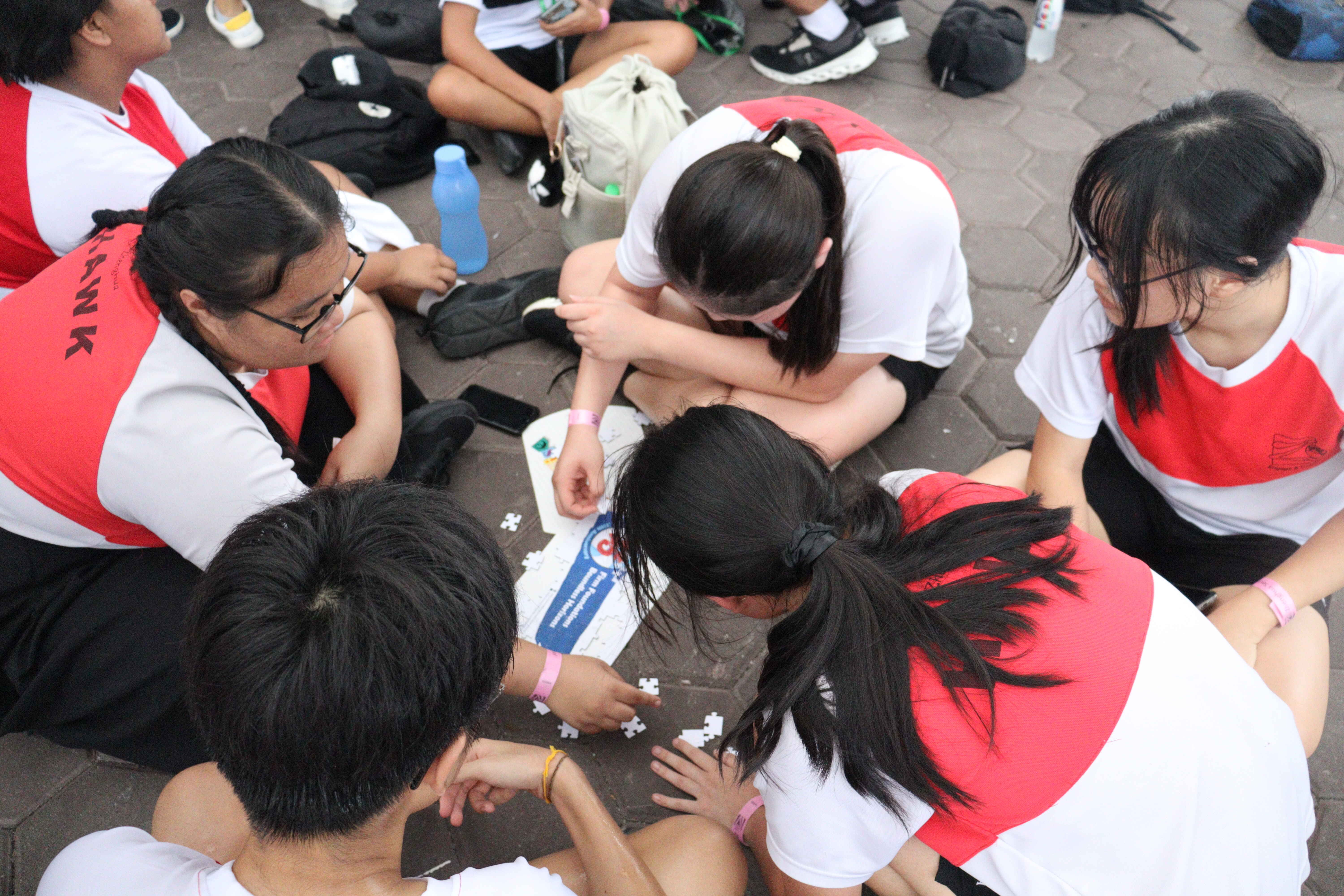 Several students in red and white uniforms sit on pavement, working together on a jigsaw puzzle.