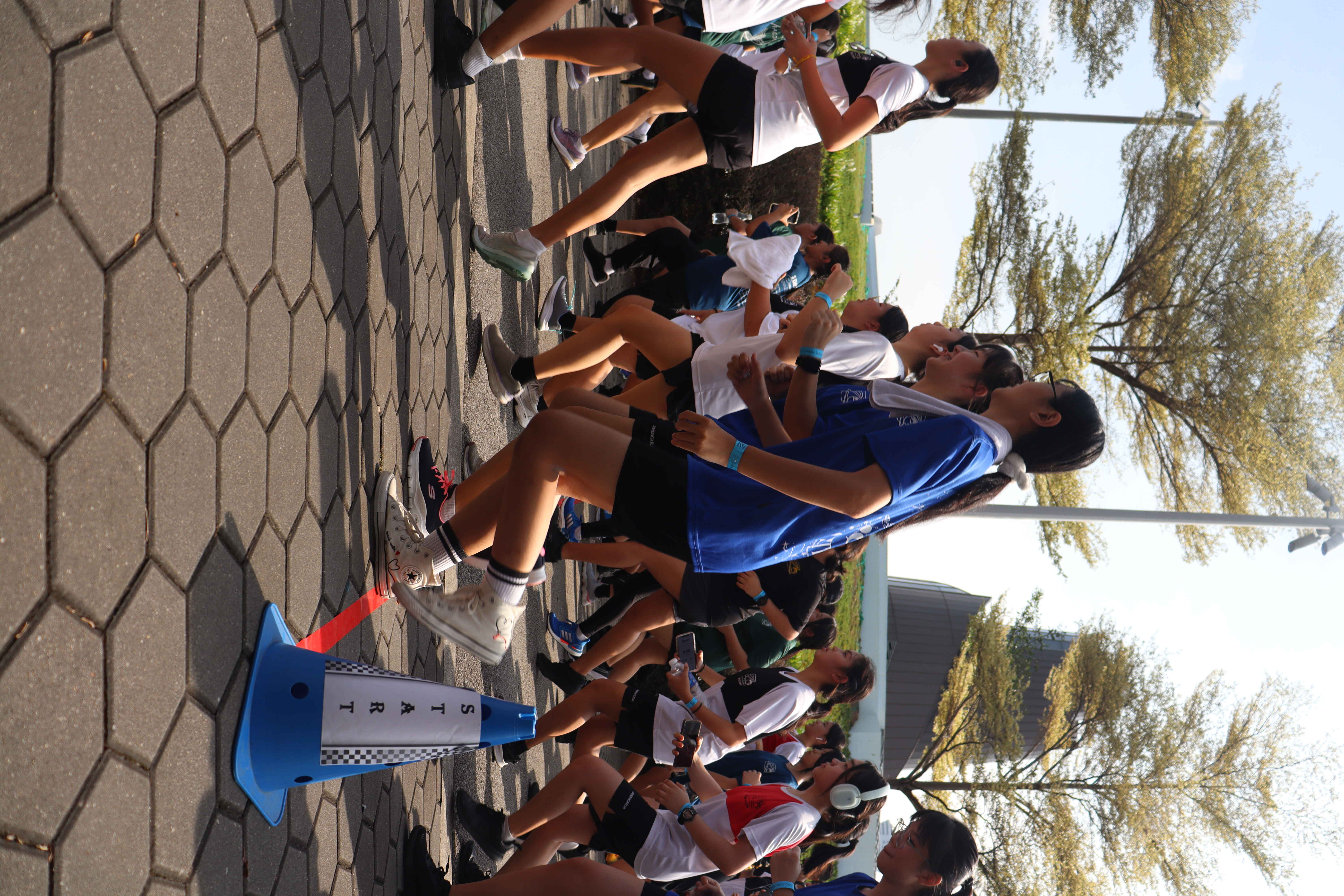 A large group of young runners crossing a blue START cone marker on a paved outdoor track surrounded by trees.