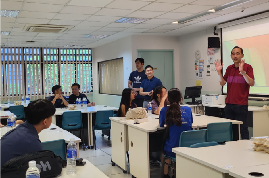 A man in a red shirt gestures while speaking to students seated at desks in a classroom with a projector screen.