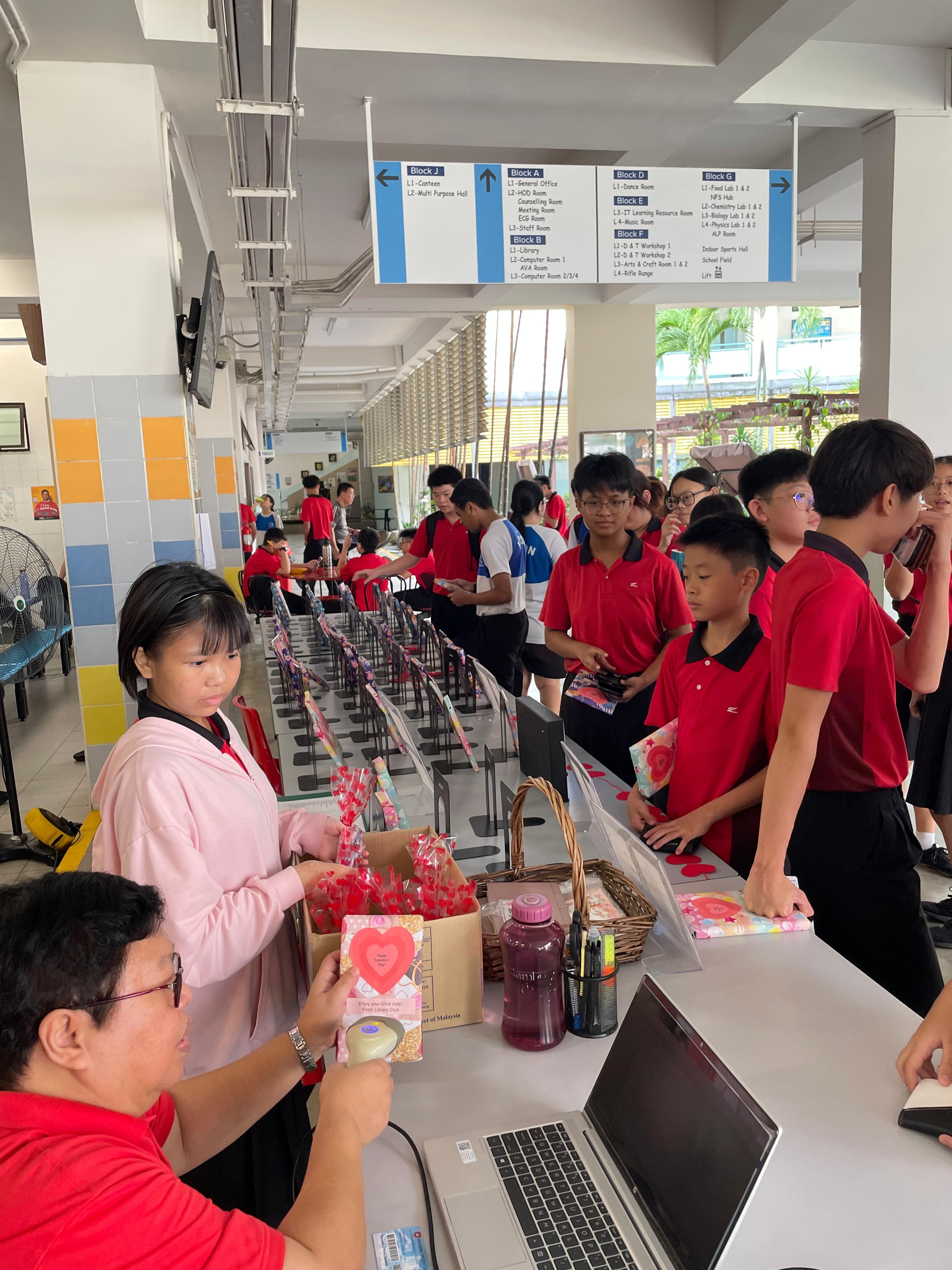 Students in red uniforms line up at a school canteen counter with candy and Valentine's goods on display.