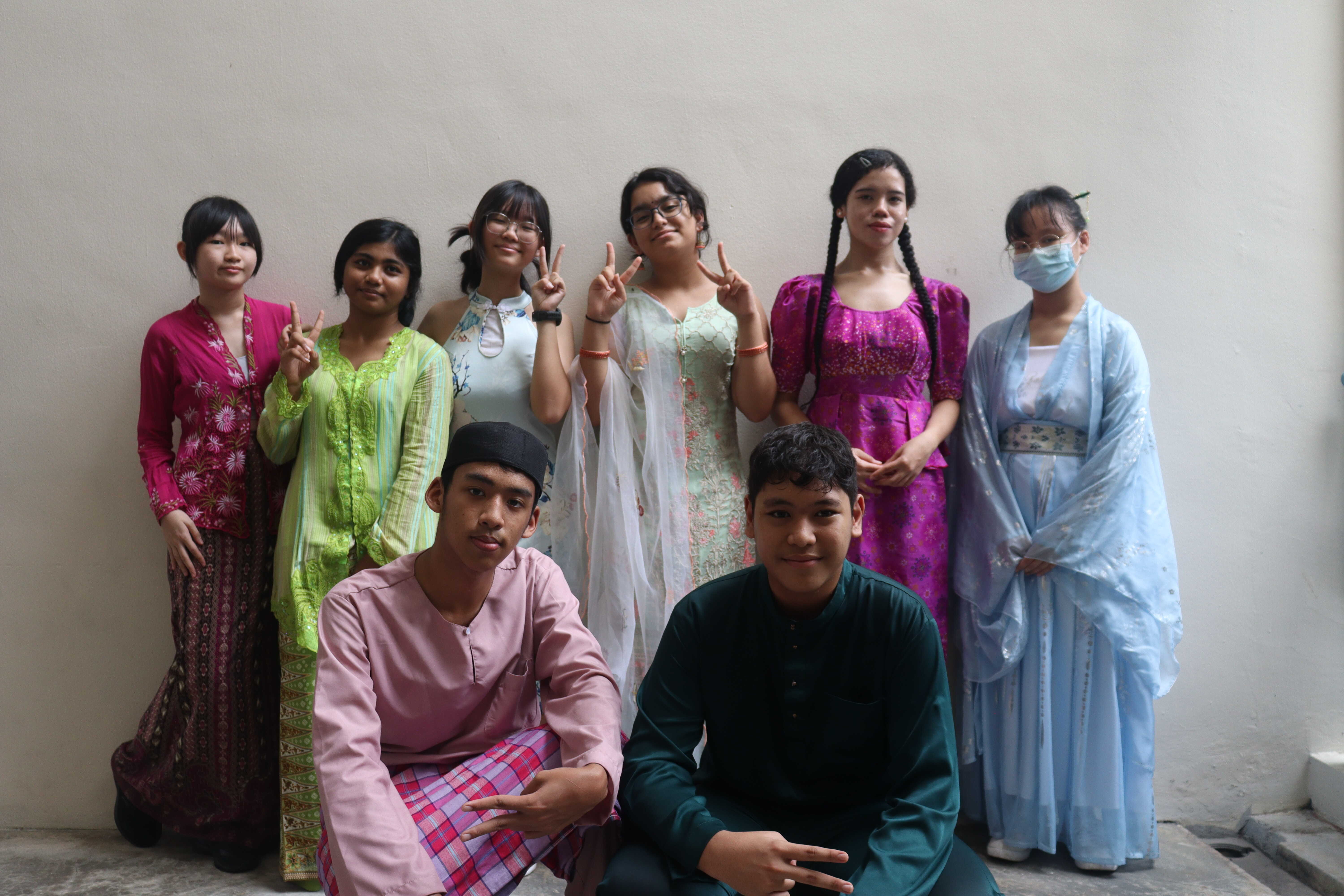 Eight young people pose in colorful traditional Southeast Asian and East Asian attire, with some making peace signs.