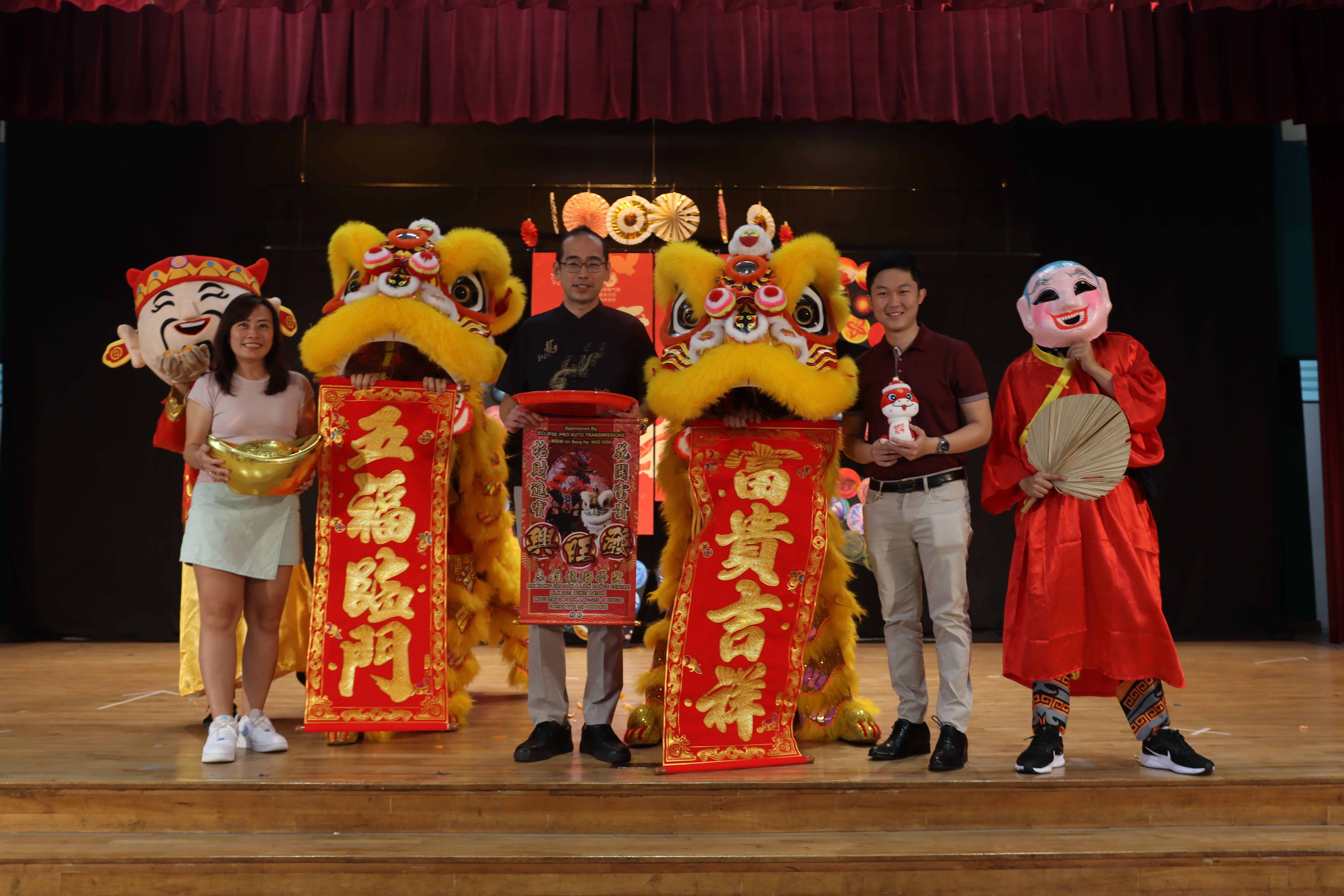 Four people on stage hold red Chinese New Year banners beside two yellow lion dance costumes and a costumed character.
