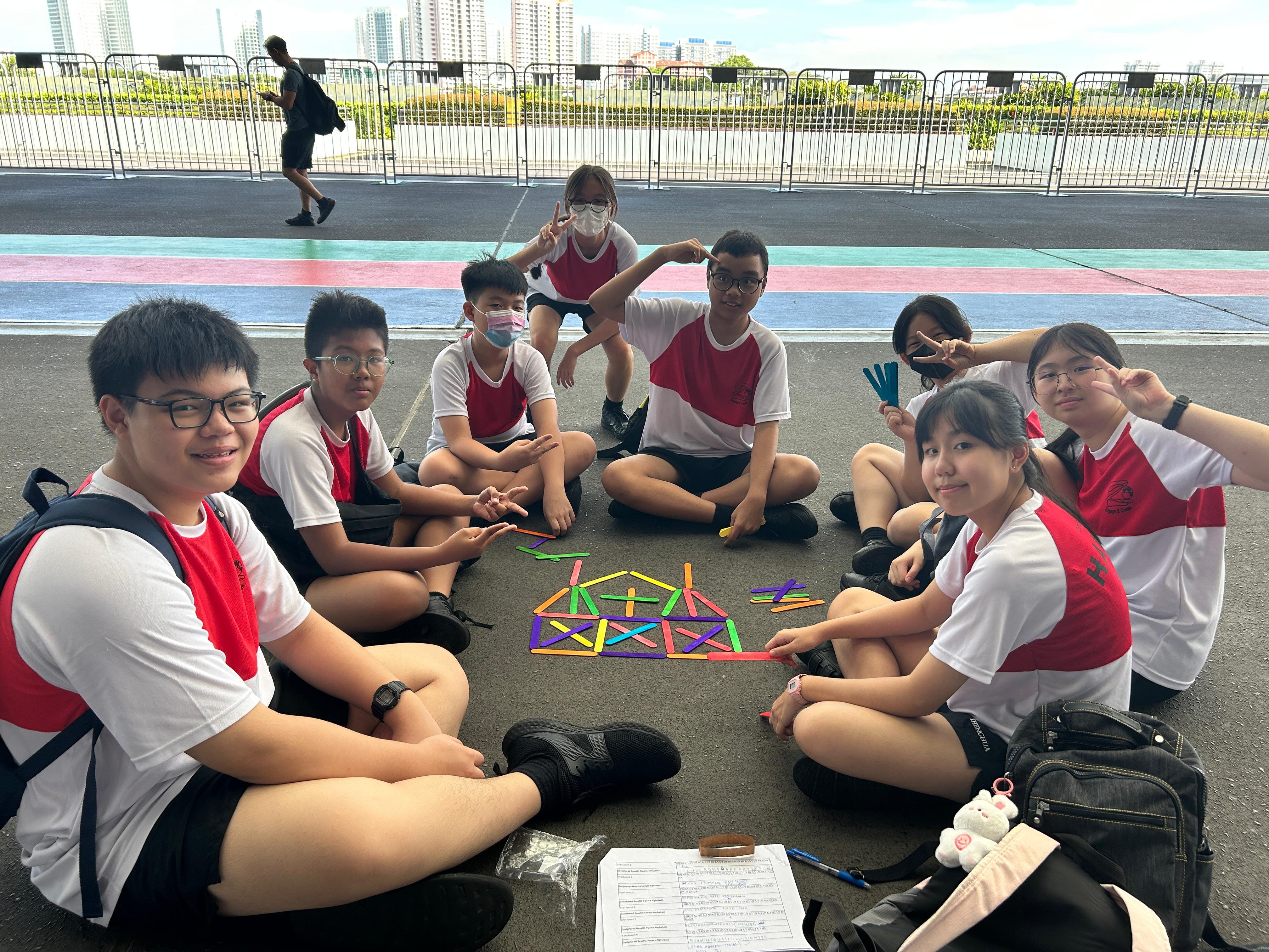 Eight students in school uniforms sit on pavement around colorful craft sticks arranged in geometric shapes.