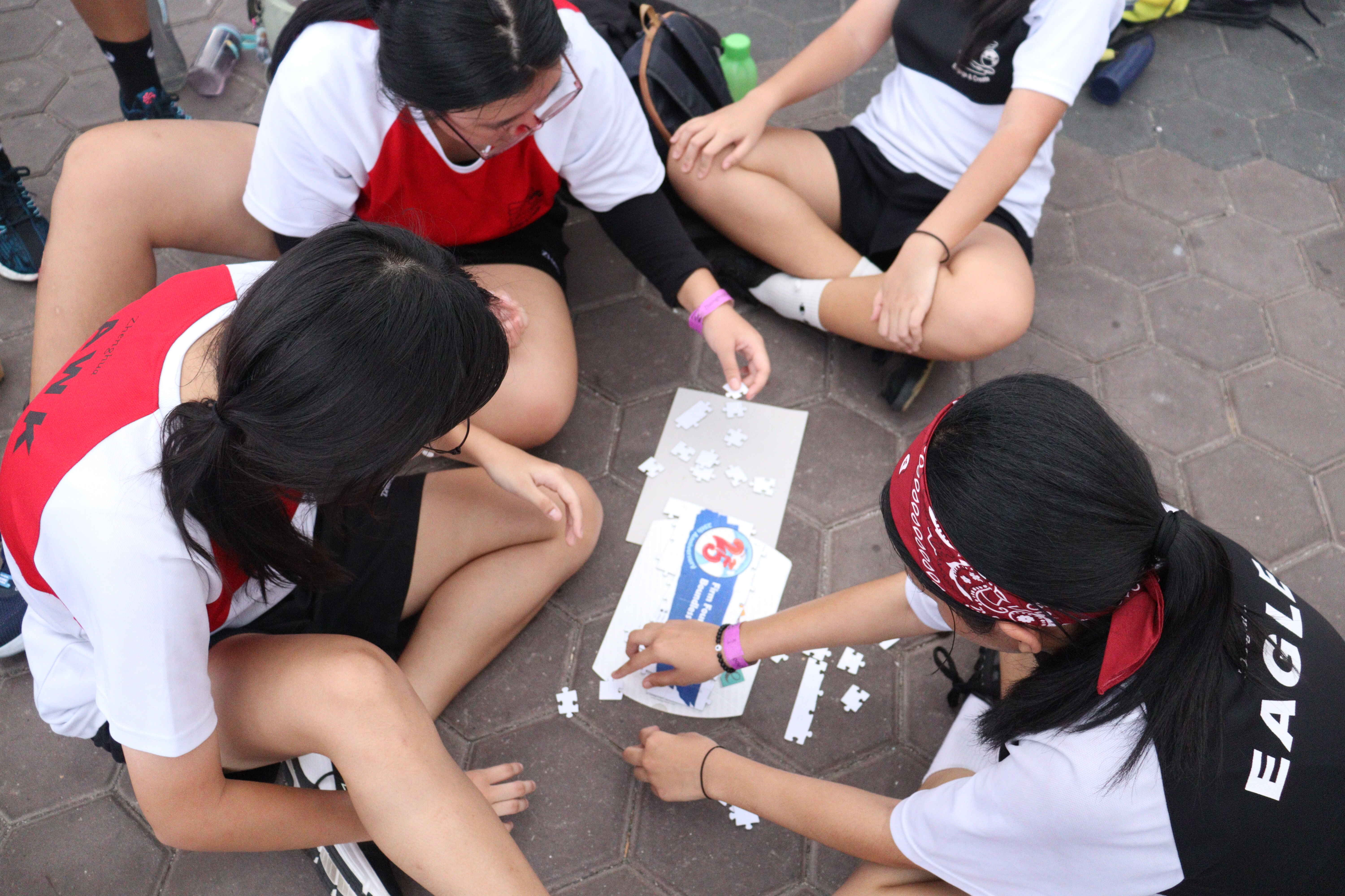 Four girls in uniforms sit on paved ground assembling a jigsaw puzzle together outdoors.