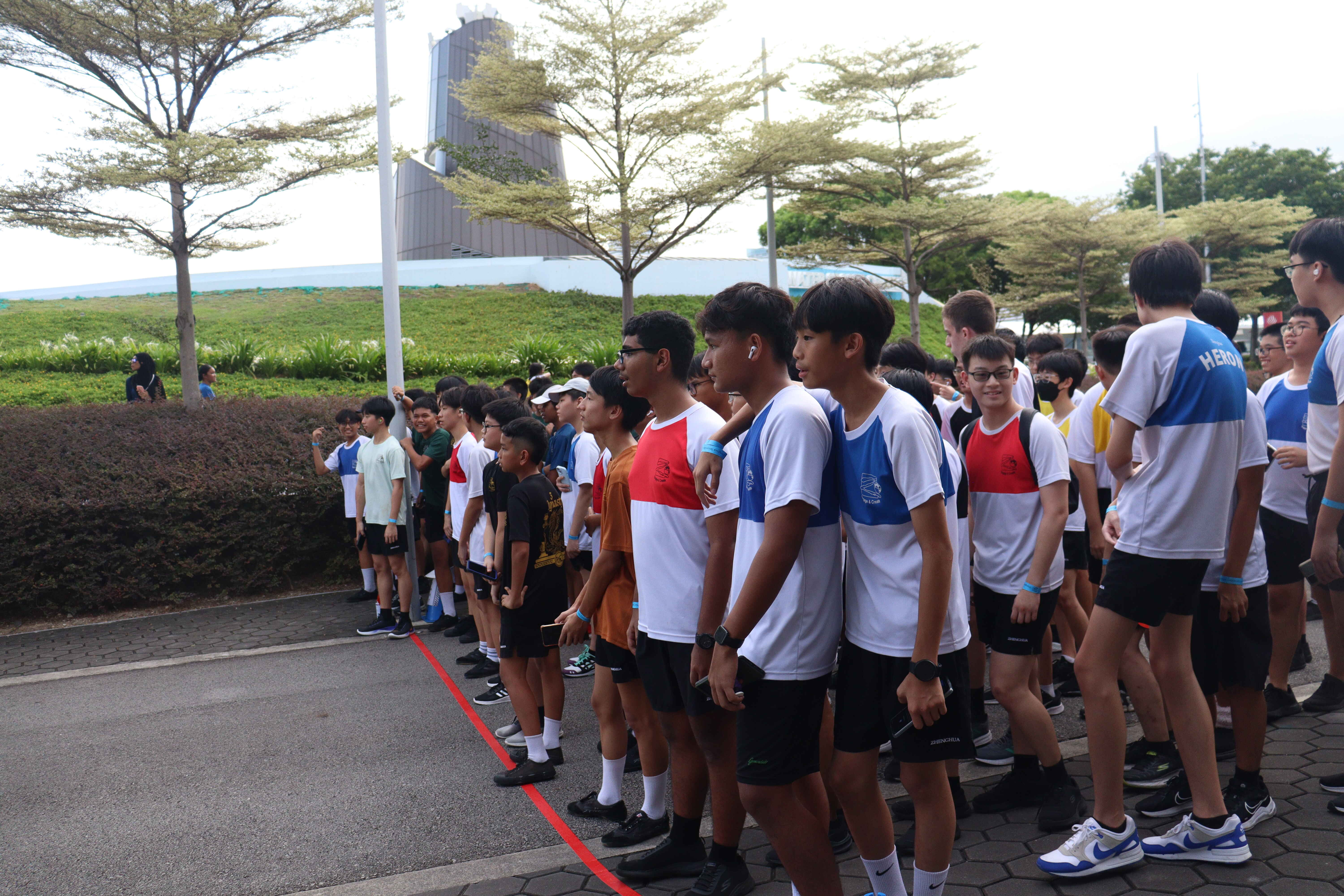 A large group of teenage boys in sports attire lined up along a red line on a paved outdoor area near trees.