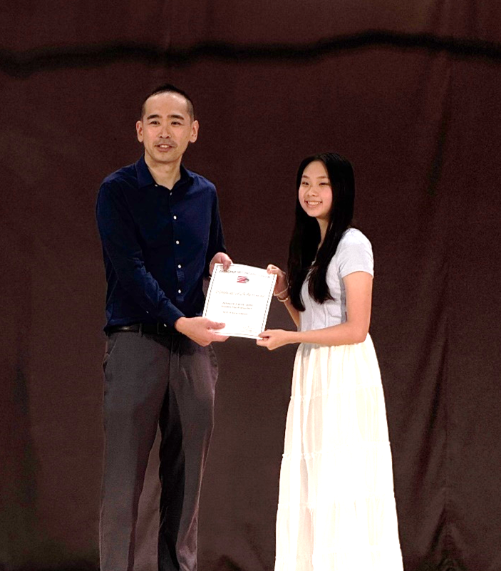 A man in a blue shirt and a young woman in a white dress hold a certificate together on a stage.