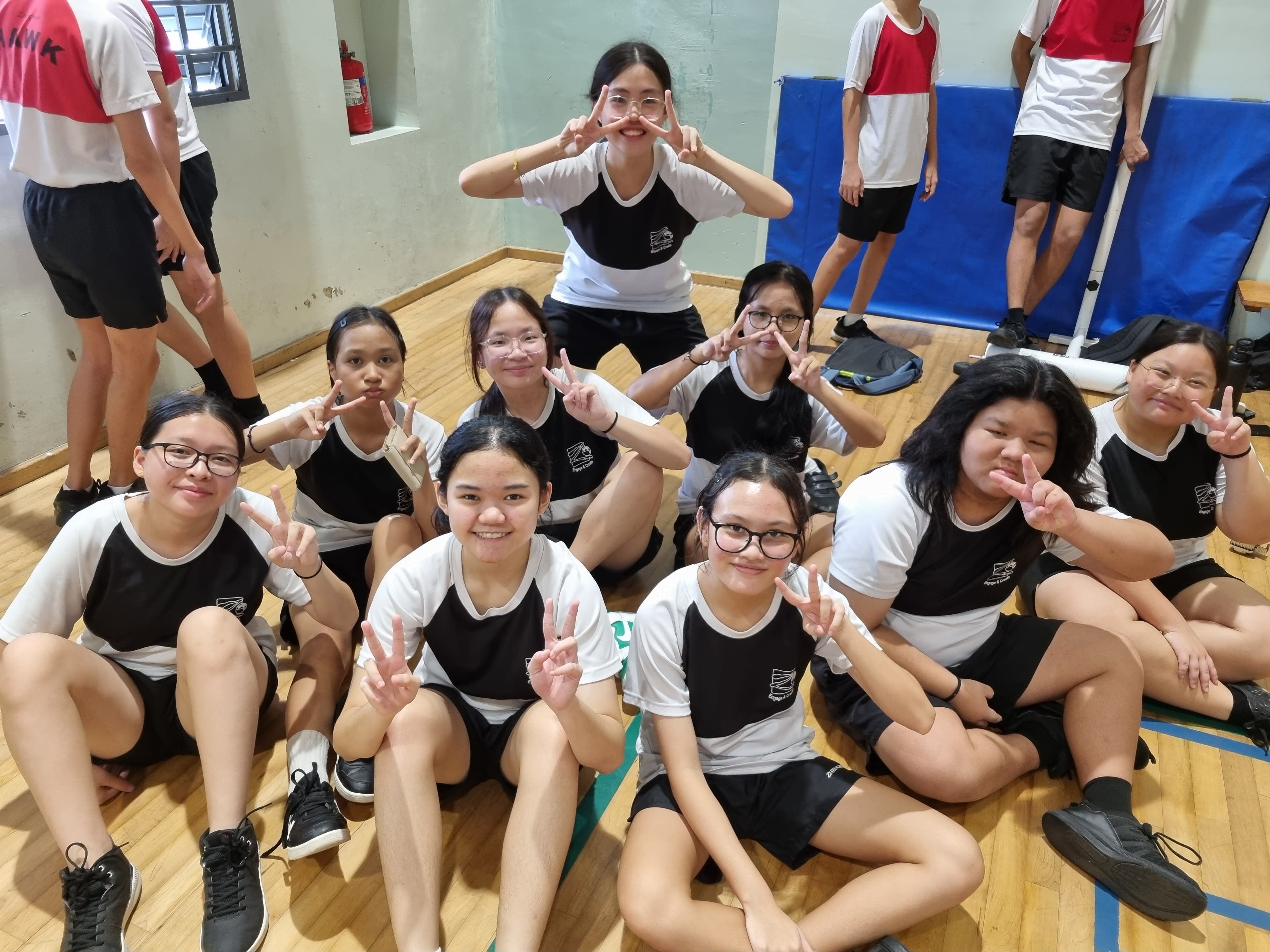 A group of young students in black and white uniforms sit on a gym floor, smiling and making peace signs.