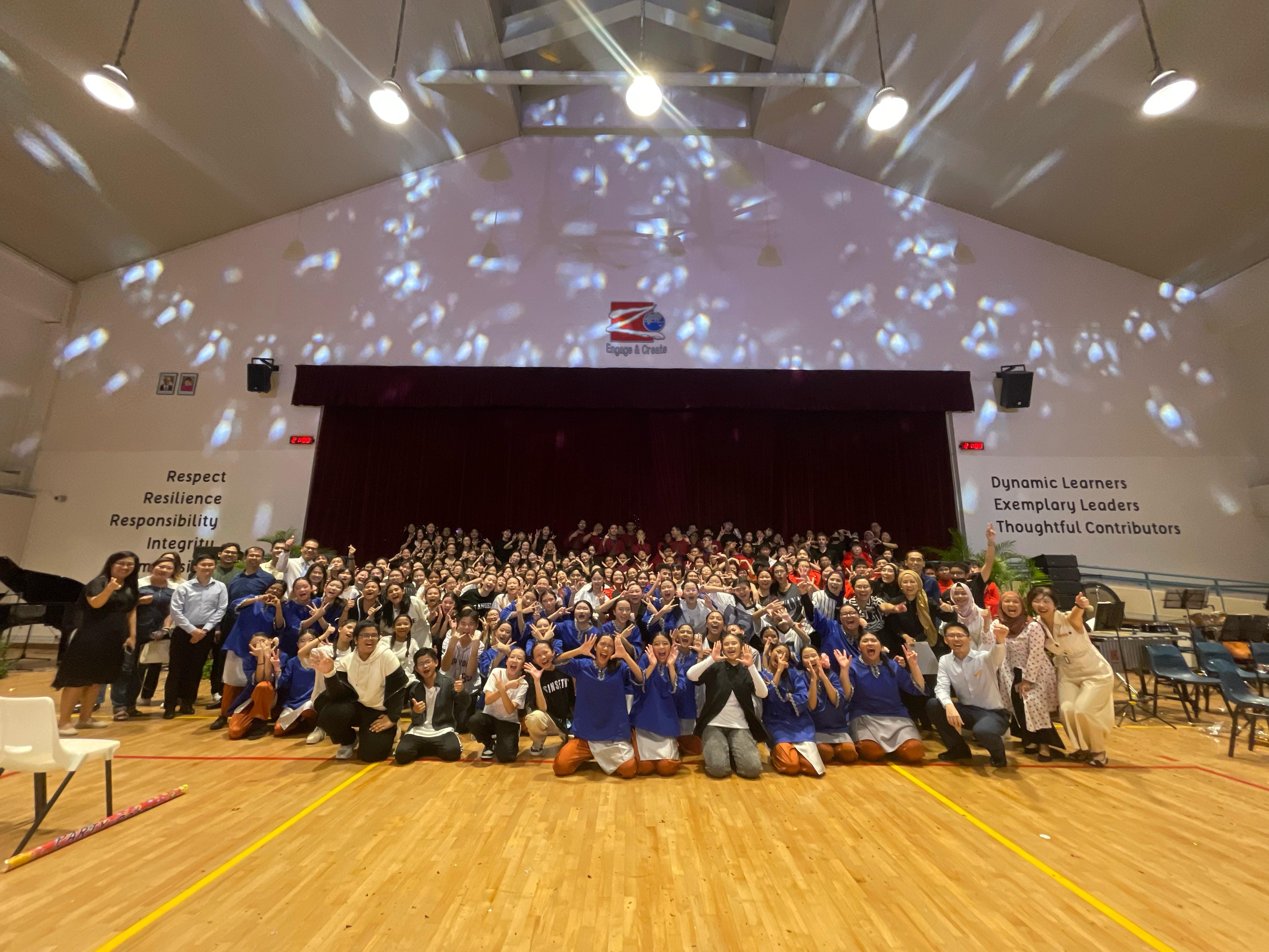 Large group of students and teachers posing in a school hall with a stage and motivational words on the walls.