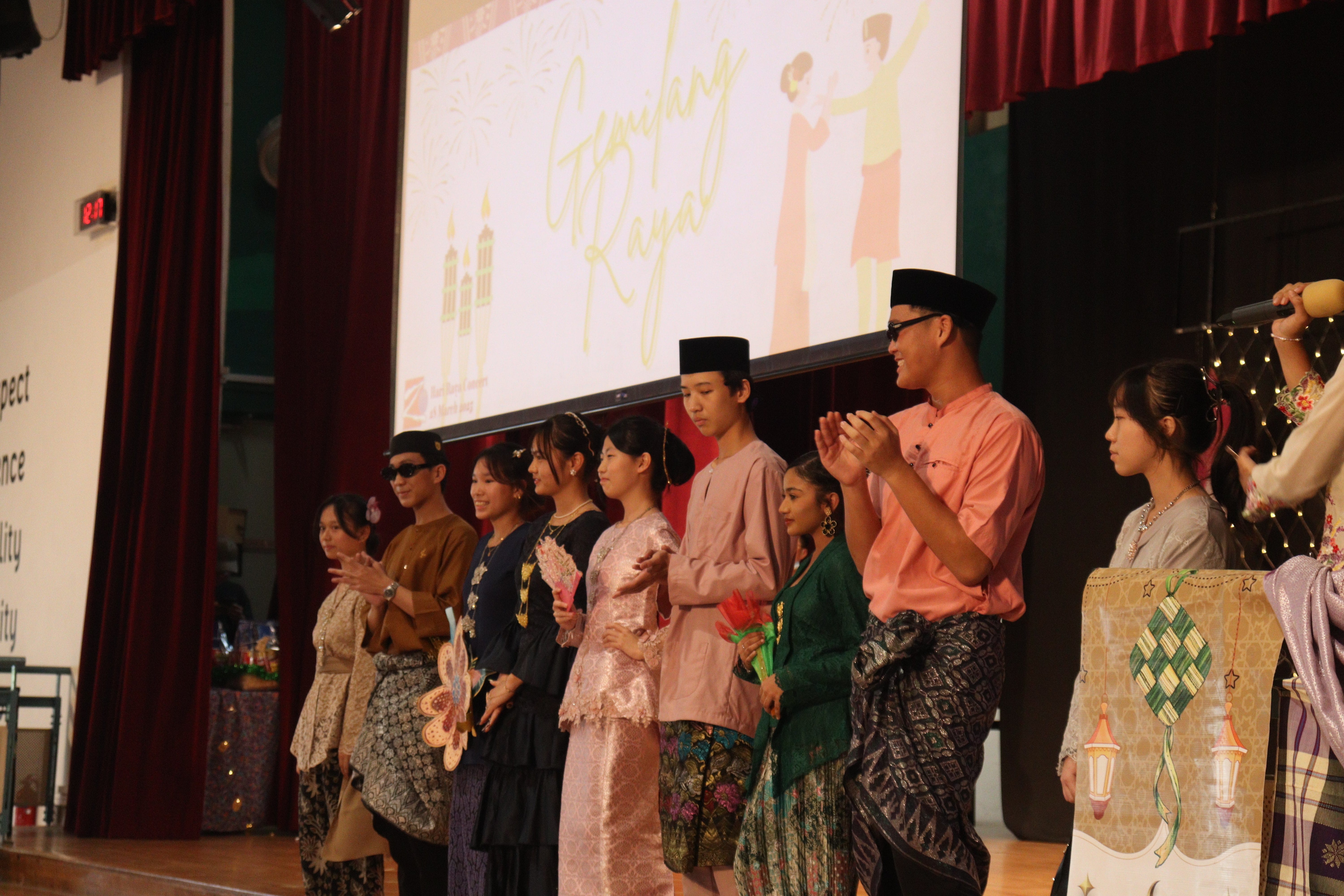 A group of young people in traditional Malay attire stand on stage at a Hari Raya school event.