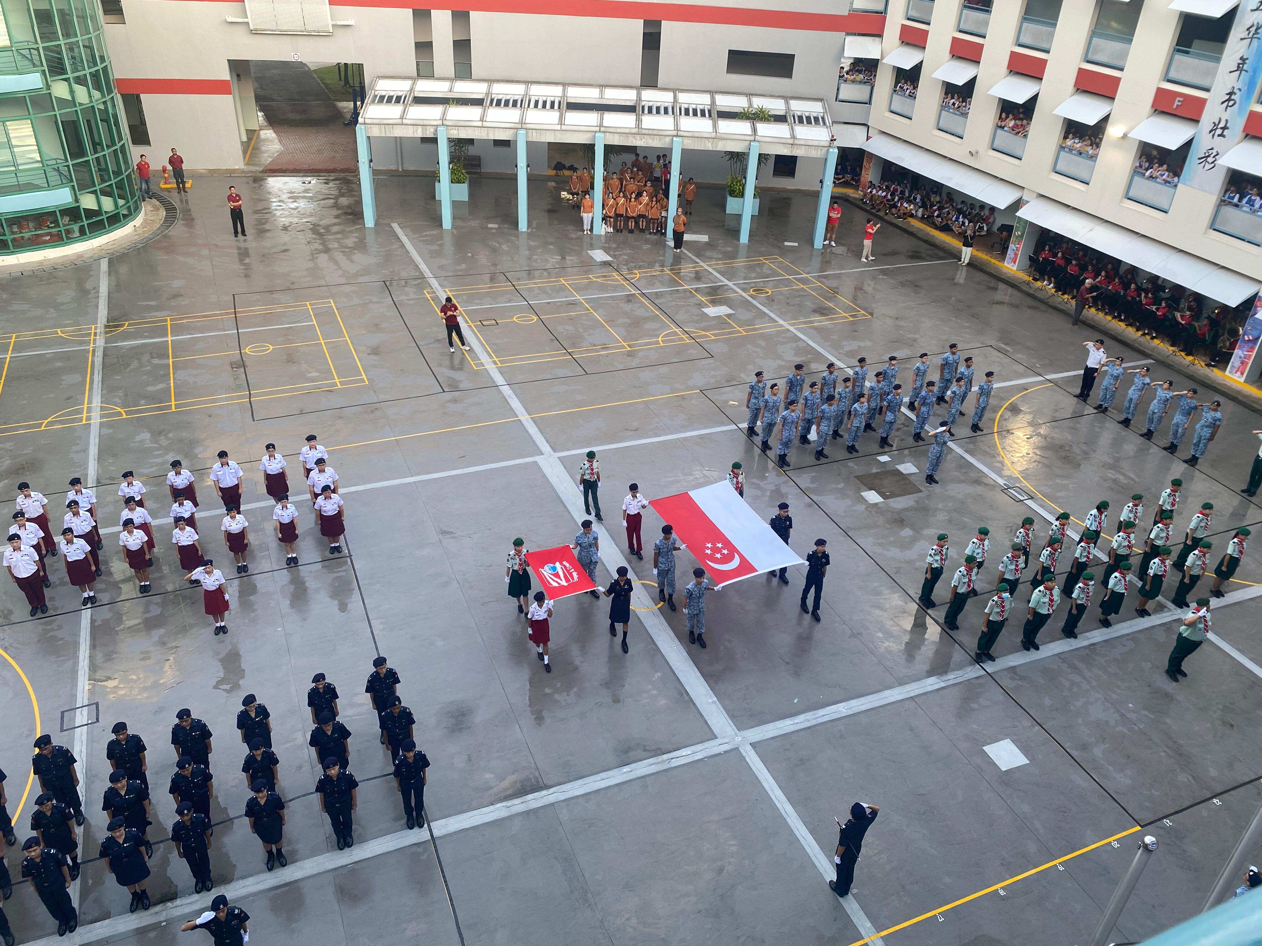 School flag ceremony on a wet courtyard with uniformed student groups standing in formation around flag bearers holding the Singapore flag.