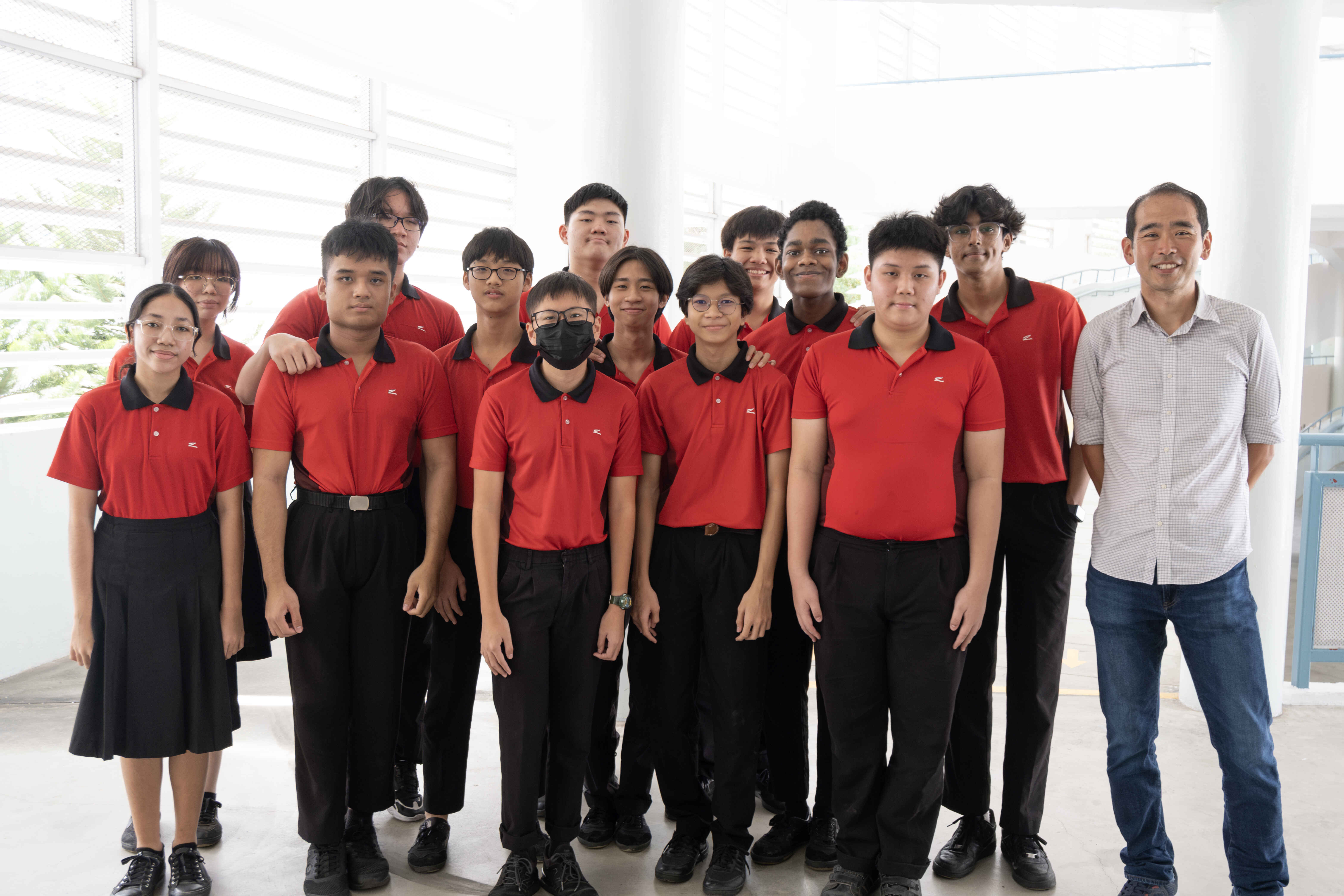 A group of students in red and black uniforms pose with an adult man in a gray shirt indoors.