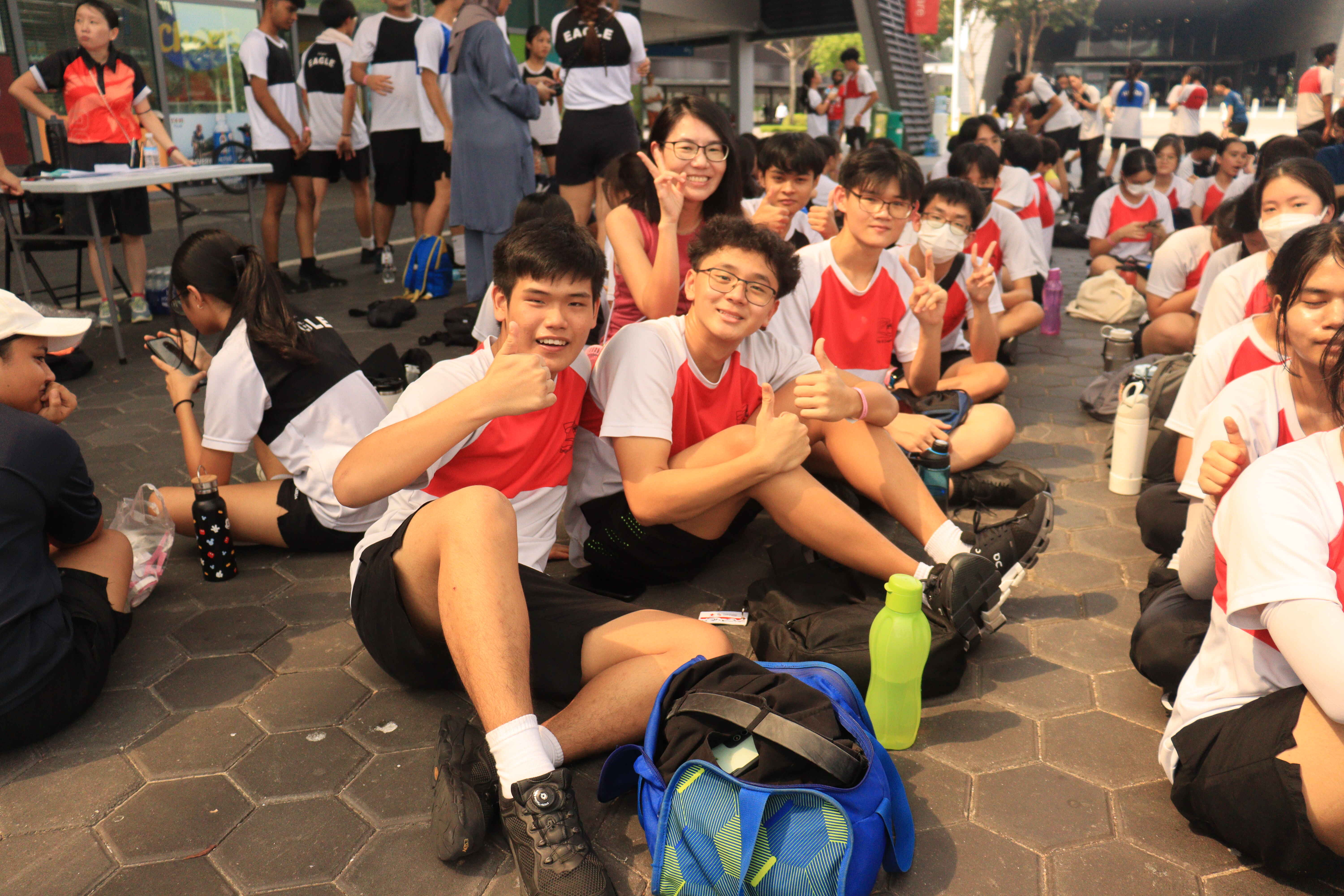 Students in red and white uniforms sit on a paved outdoor area, smiling and giving thumbs up at a school event.
