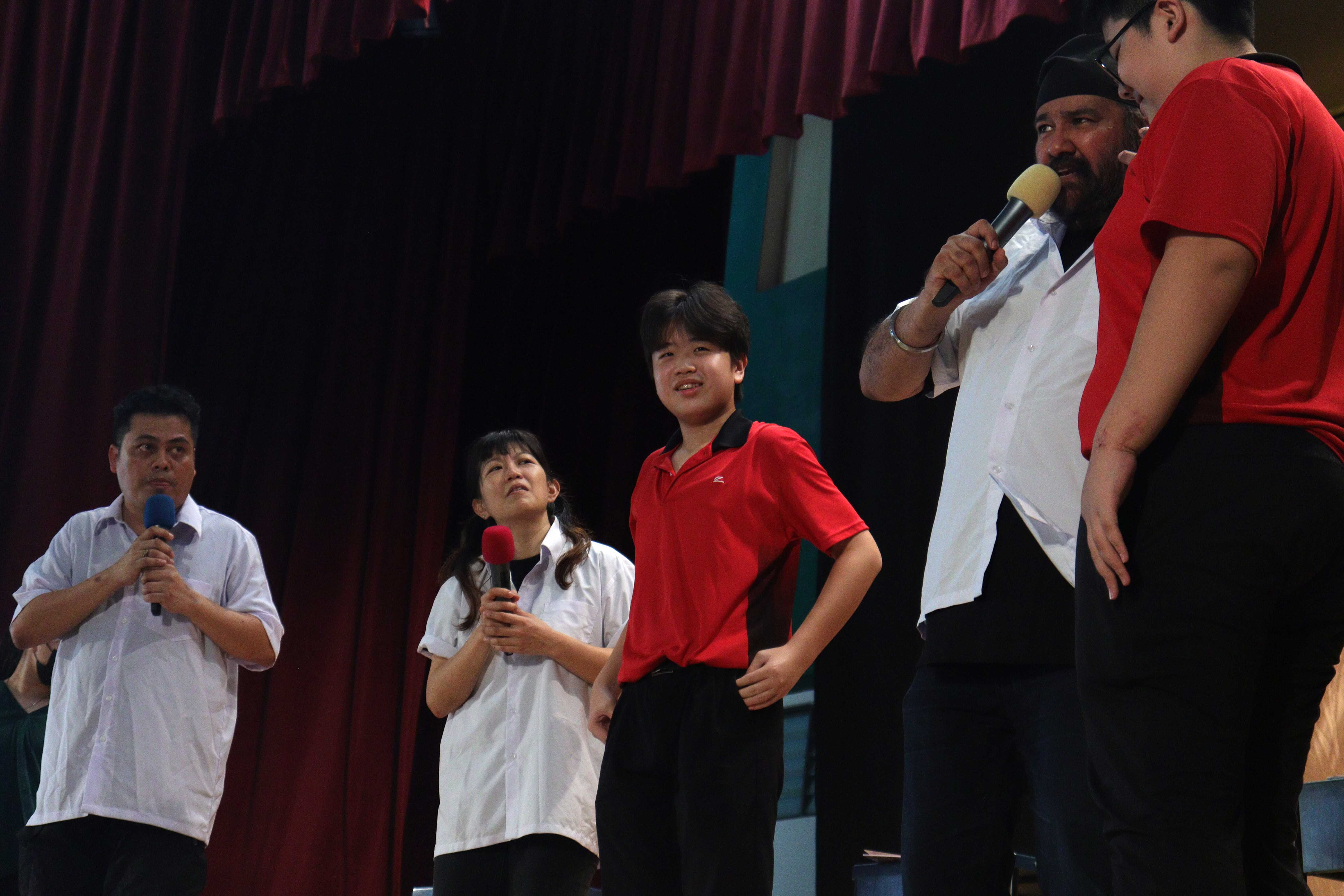 Five people stand on a stage with red curtains, some holding microphones, appearing to perform or speak.