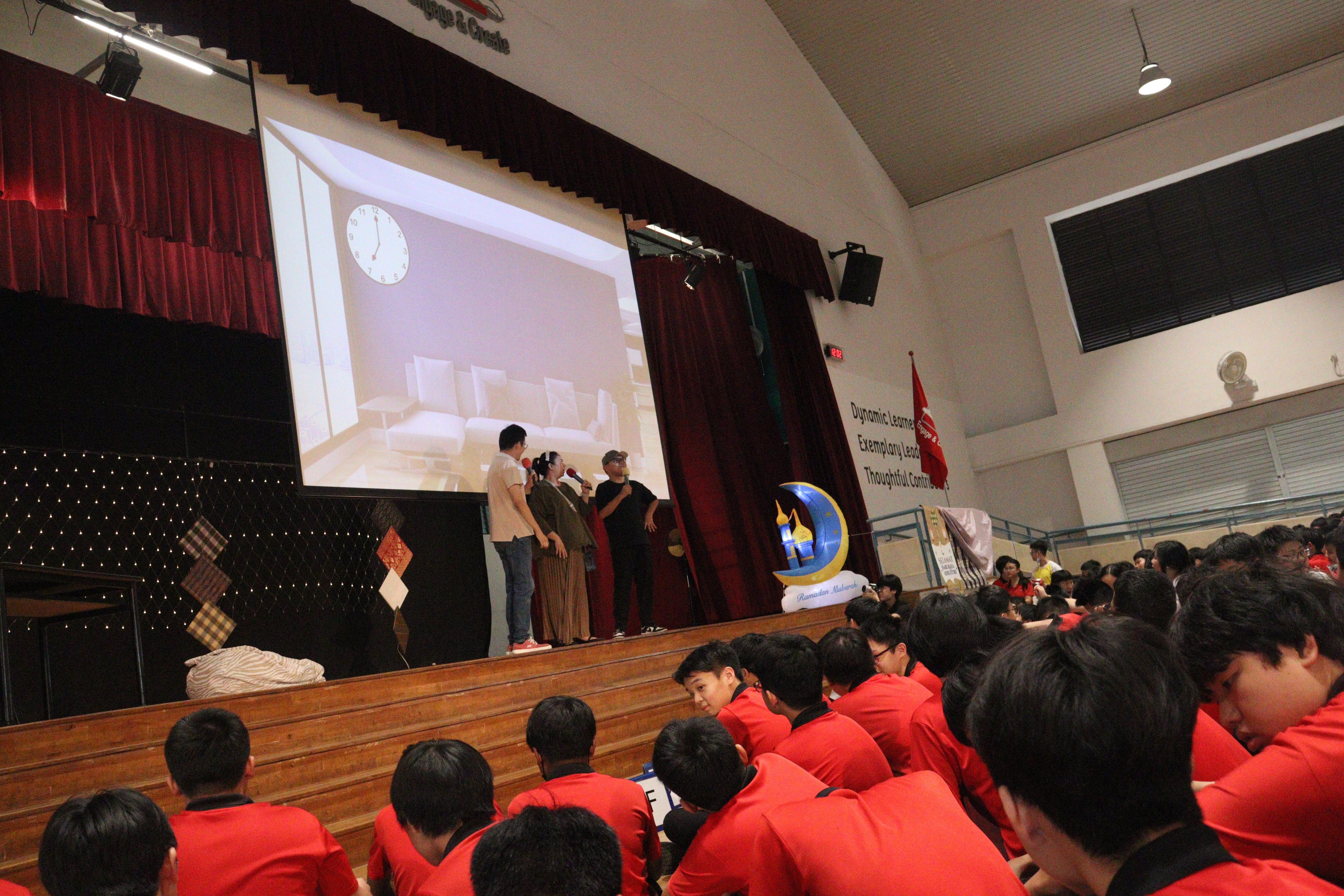 Students in red shirts sit facing a school stage where three people stand with microphones before a projected screen.