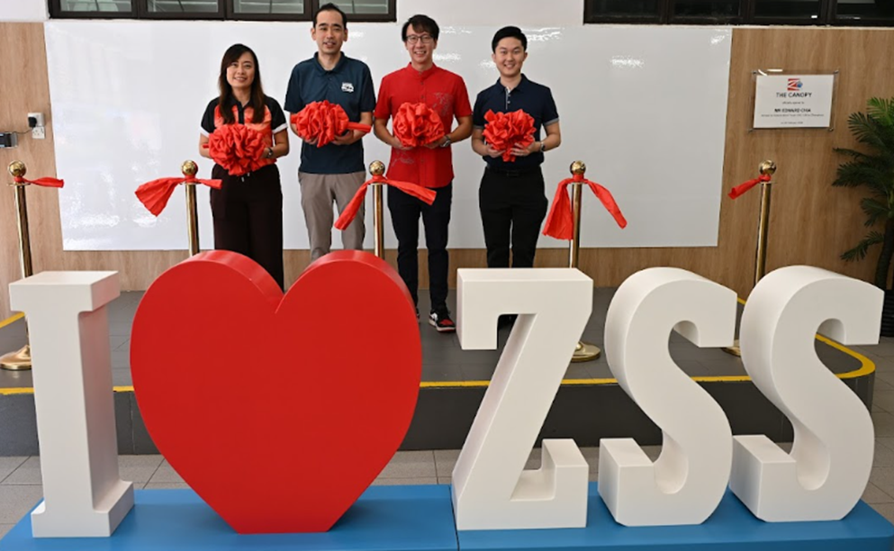 Four people hold red ribbons behind a large "I Love ZSS" sign with a red heart indoors.