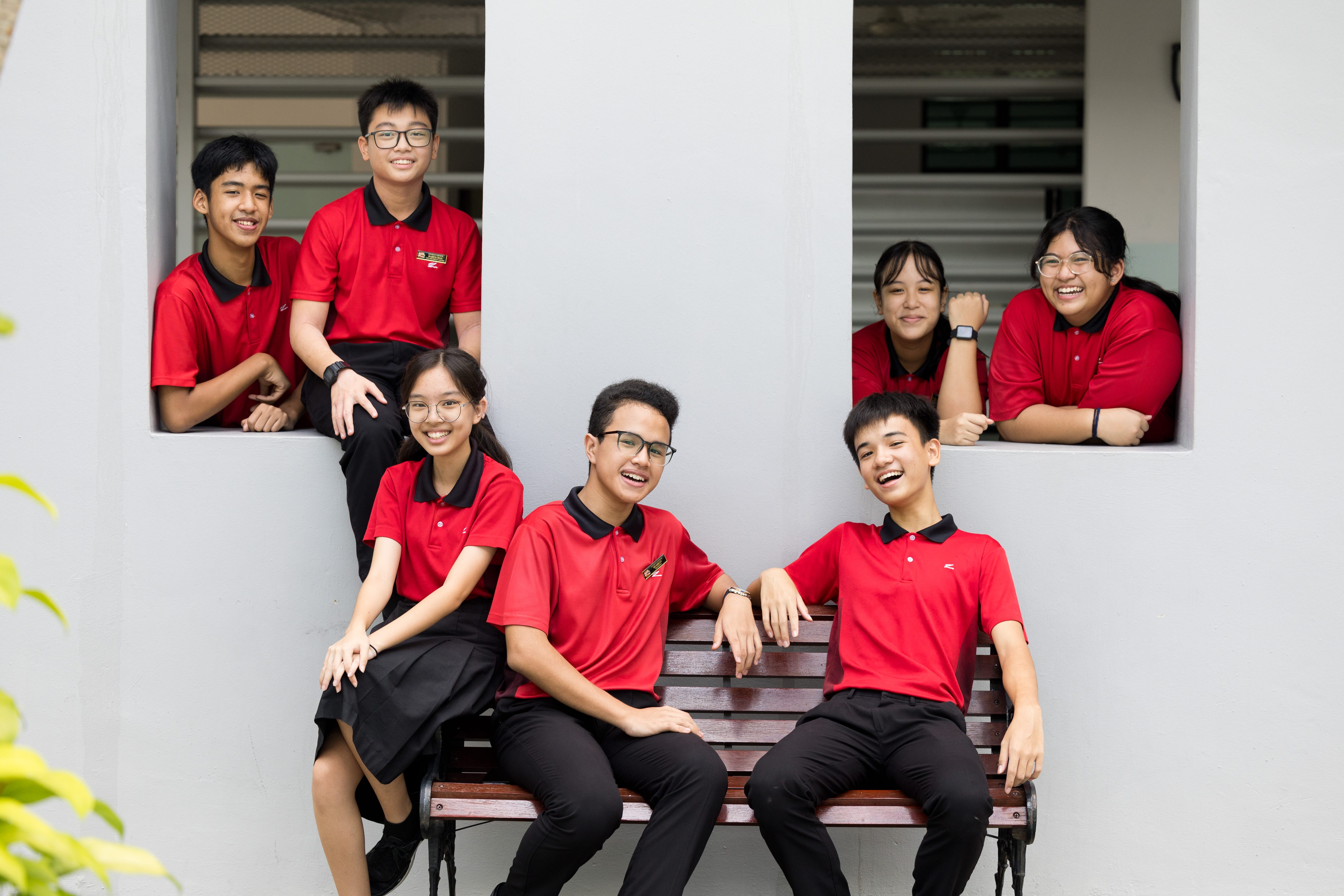 Six students in red shirts smile and pose by a window and bench against a white wall.
