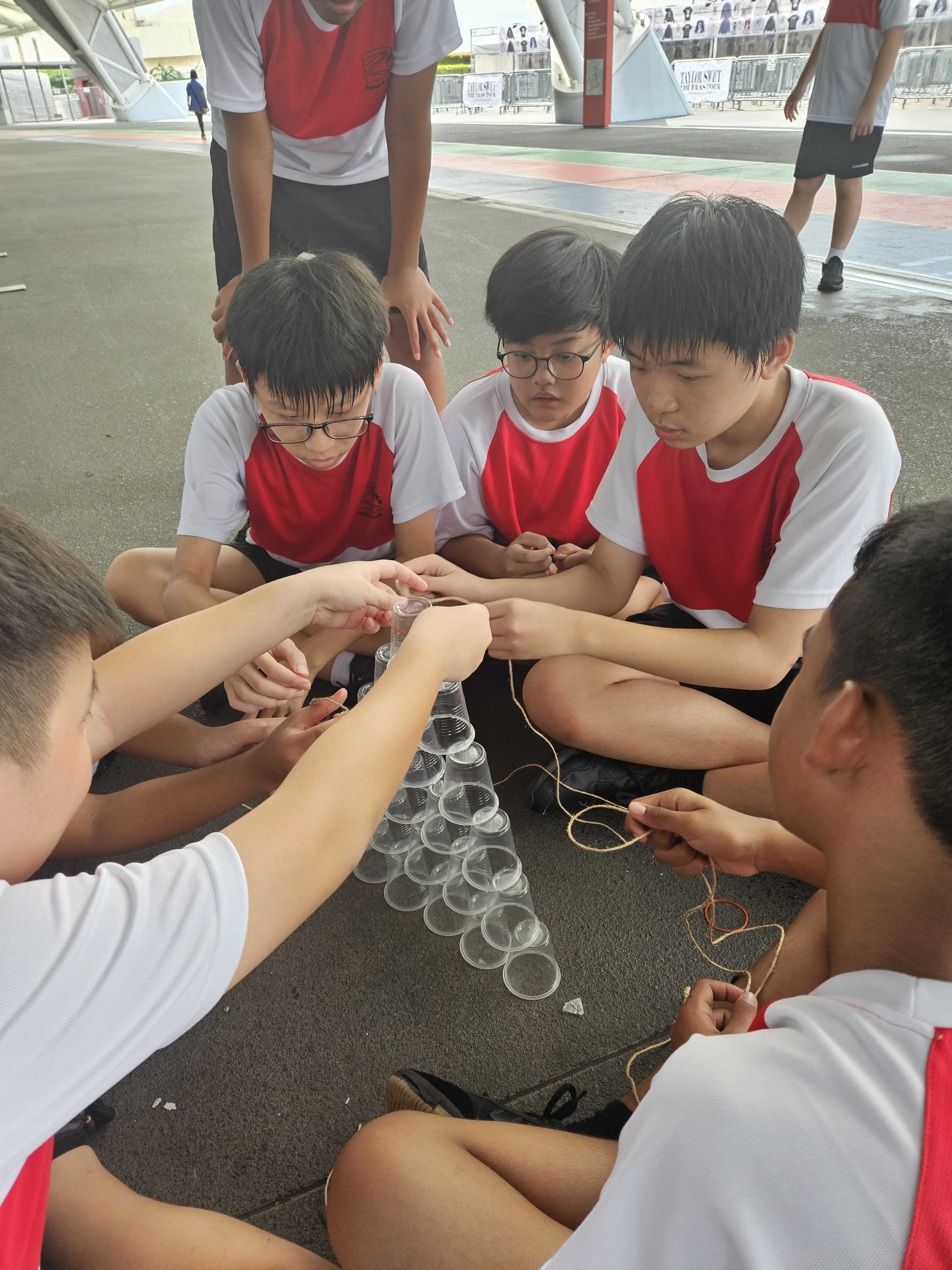 Students in red and white uniforms sit on the ground stacking clear plastic cups into a pyramid using strings.