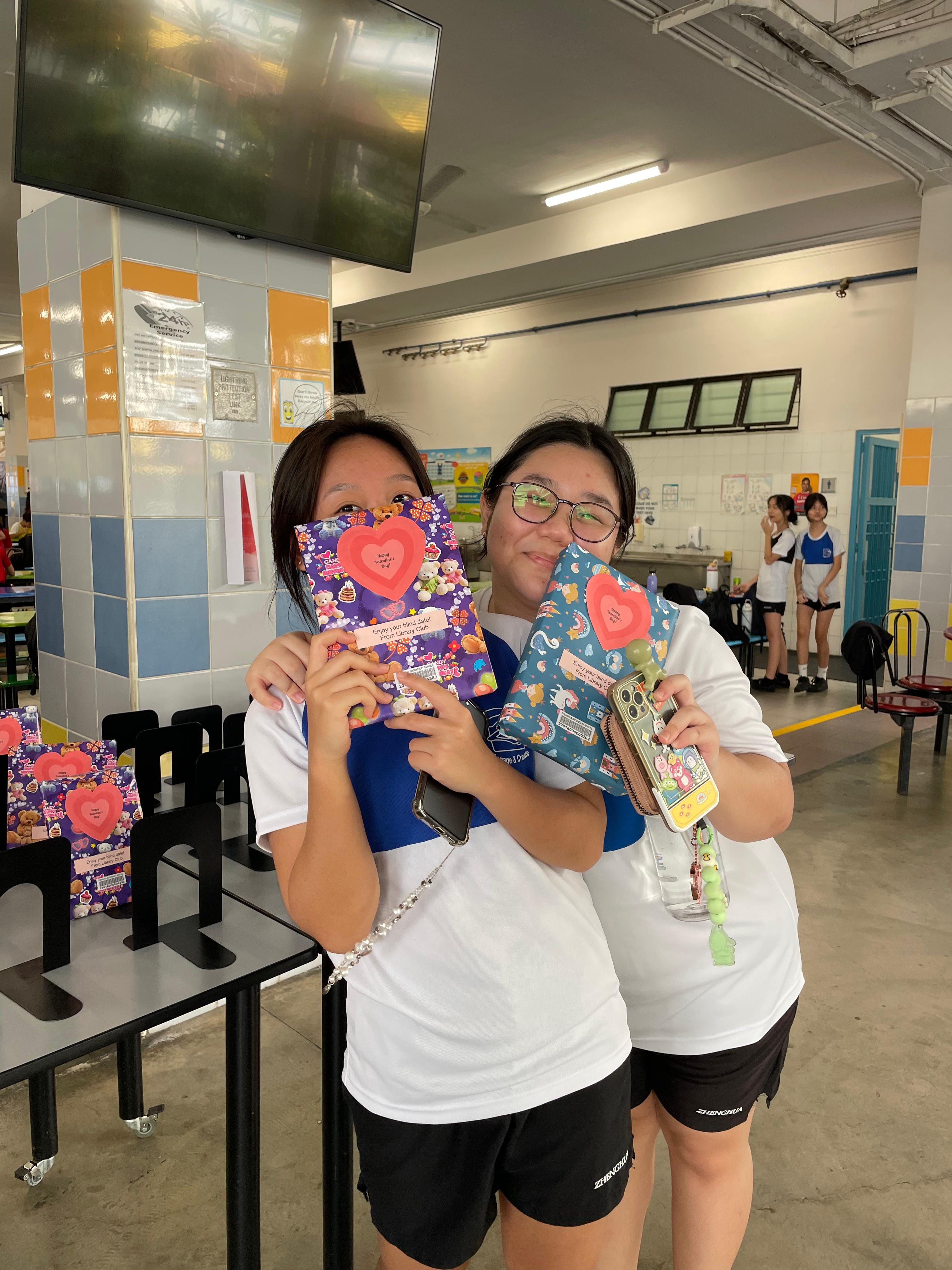 Two girls in white shirts hold up colorful wrapped books with heart decorations in a school canteen.