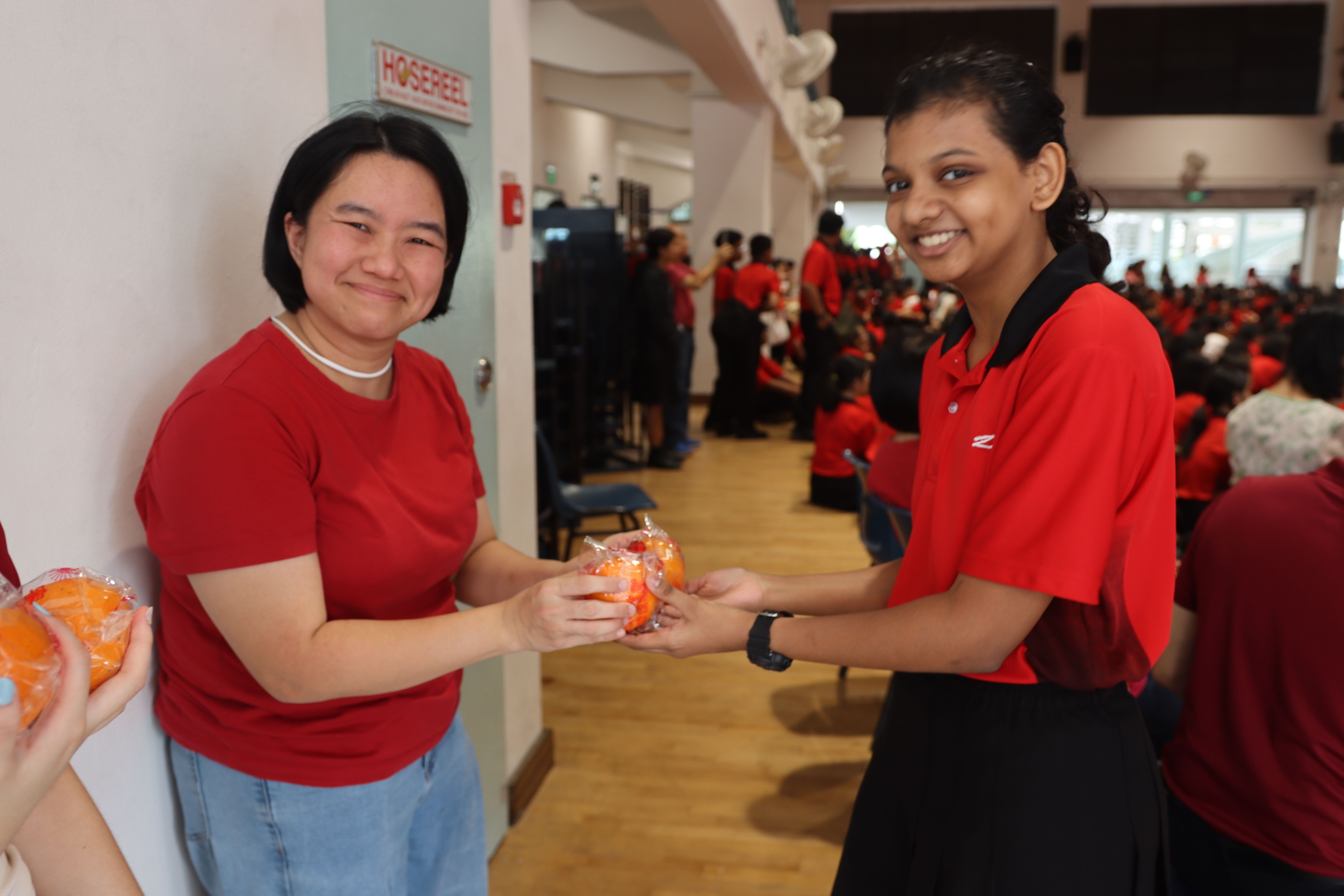Two smiling girls in red shirts exchange a wrapped orange fruit in a crowded hall filled with people in red.