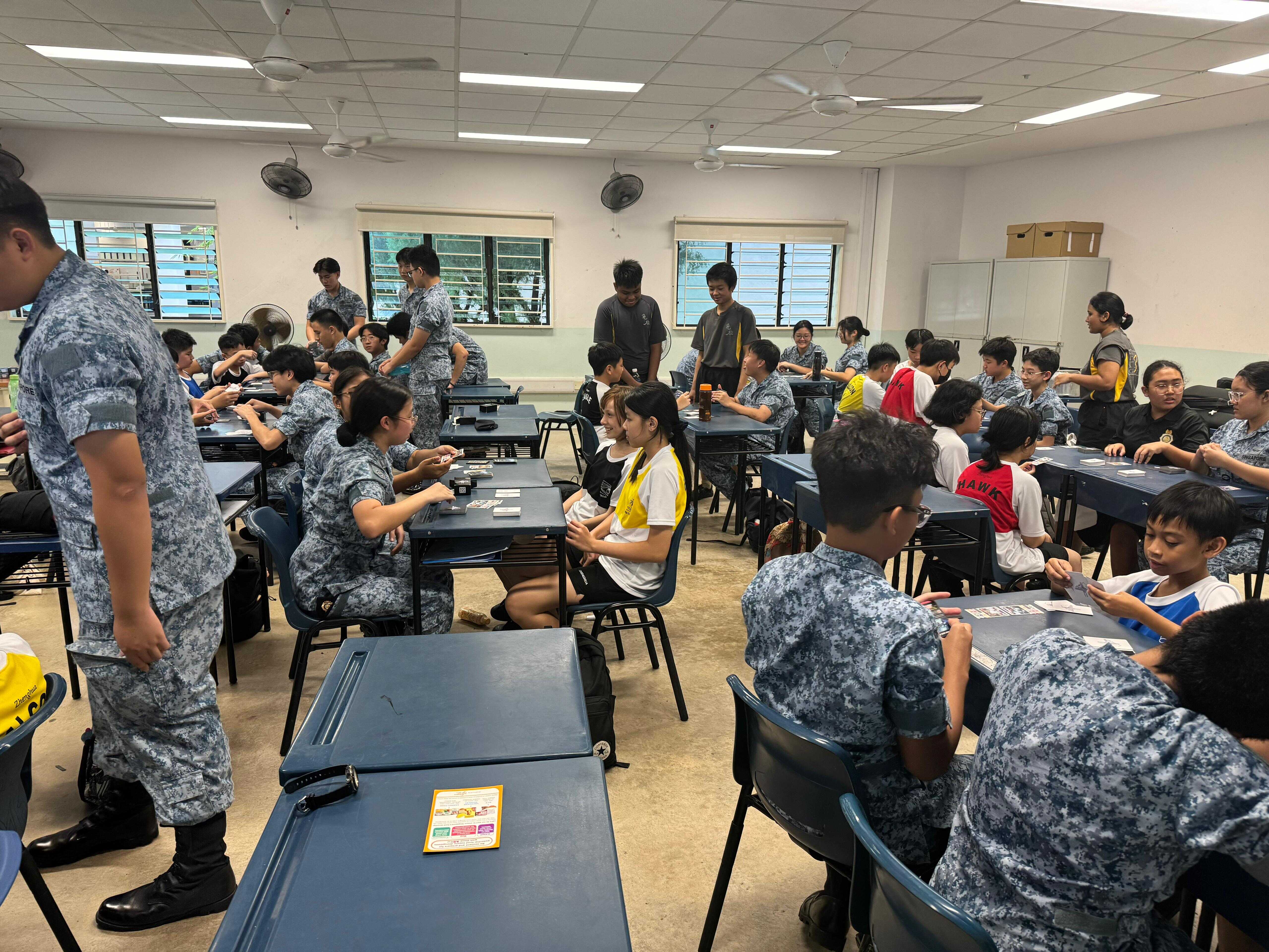 Students in blue camouflage uniforms sit at desks in a classroom, engaged in a card activity with supervisors nearby.