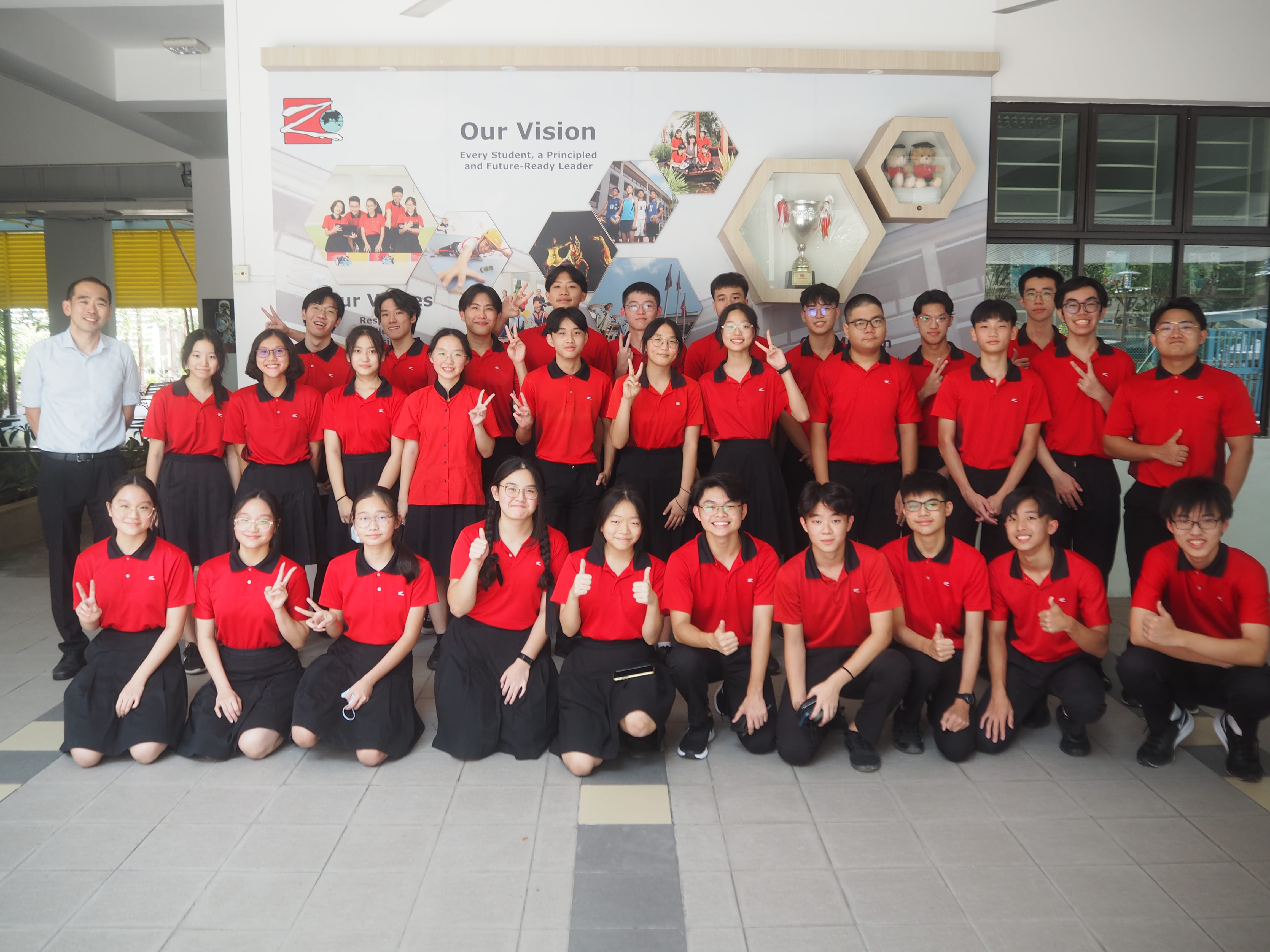A large group of students in red and black uniforms pose in front of a school "Our Vision" banner.