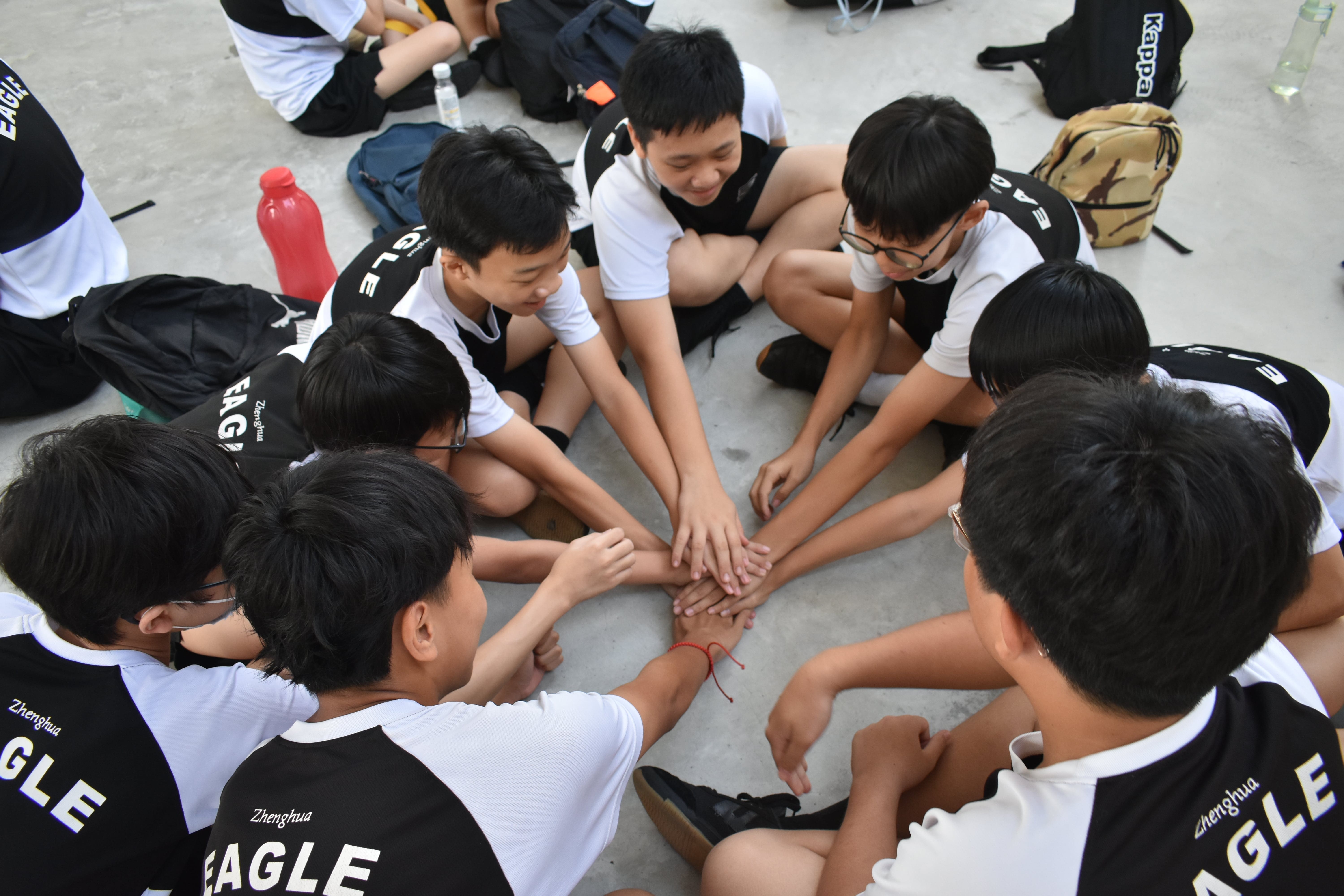 A group of boys in Zhenghua Eagle uniforms sit in a circle, stacking hands together in a team huddle.