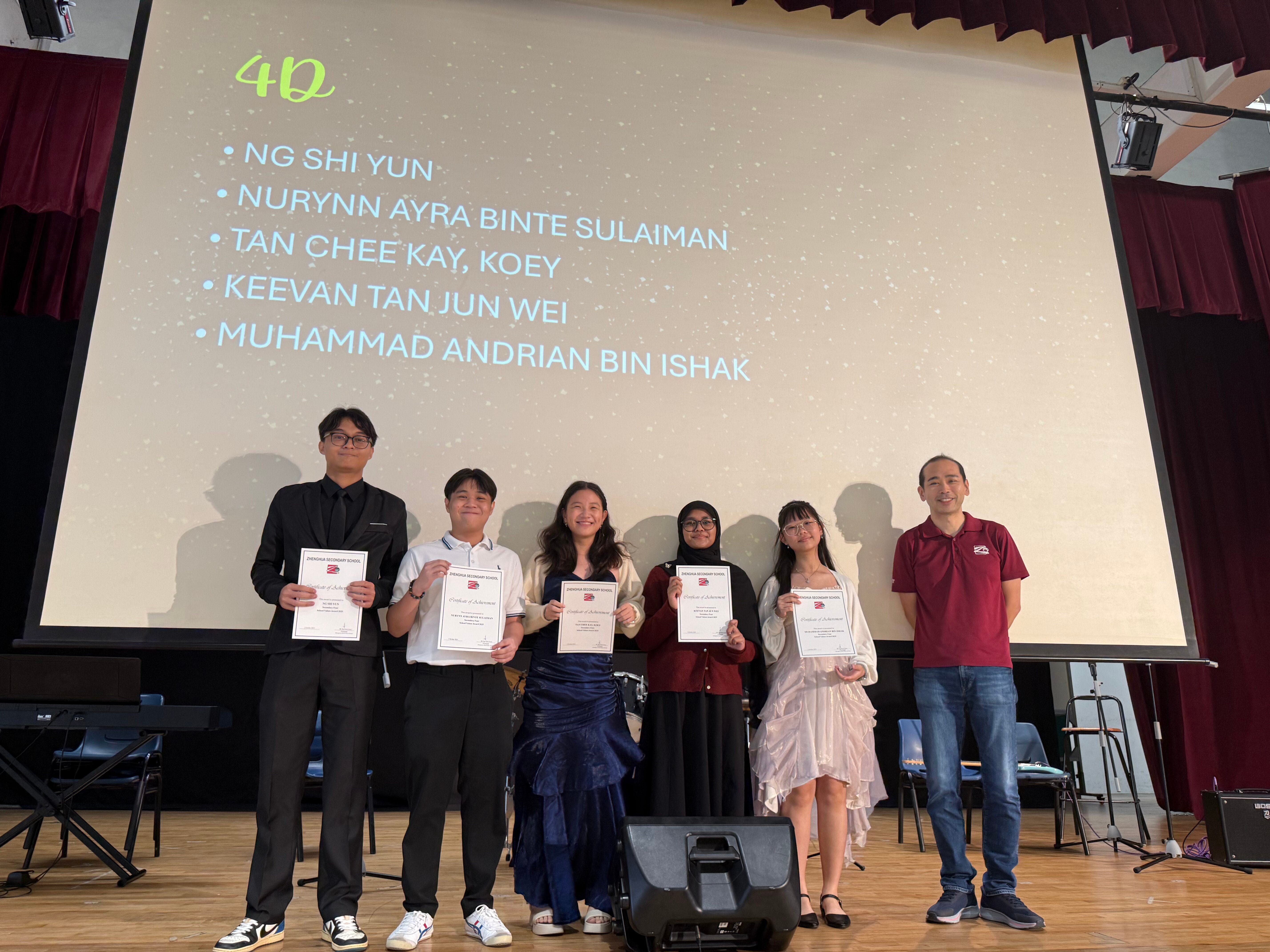 Six people stand on a stage holding certificates in front of a projected slide listing class 4D student names.