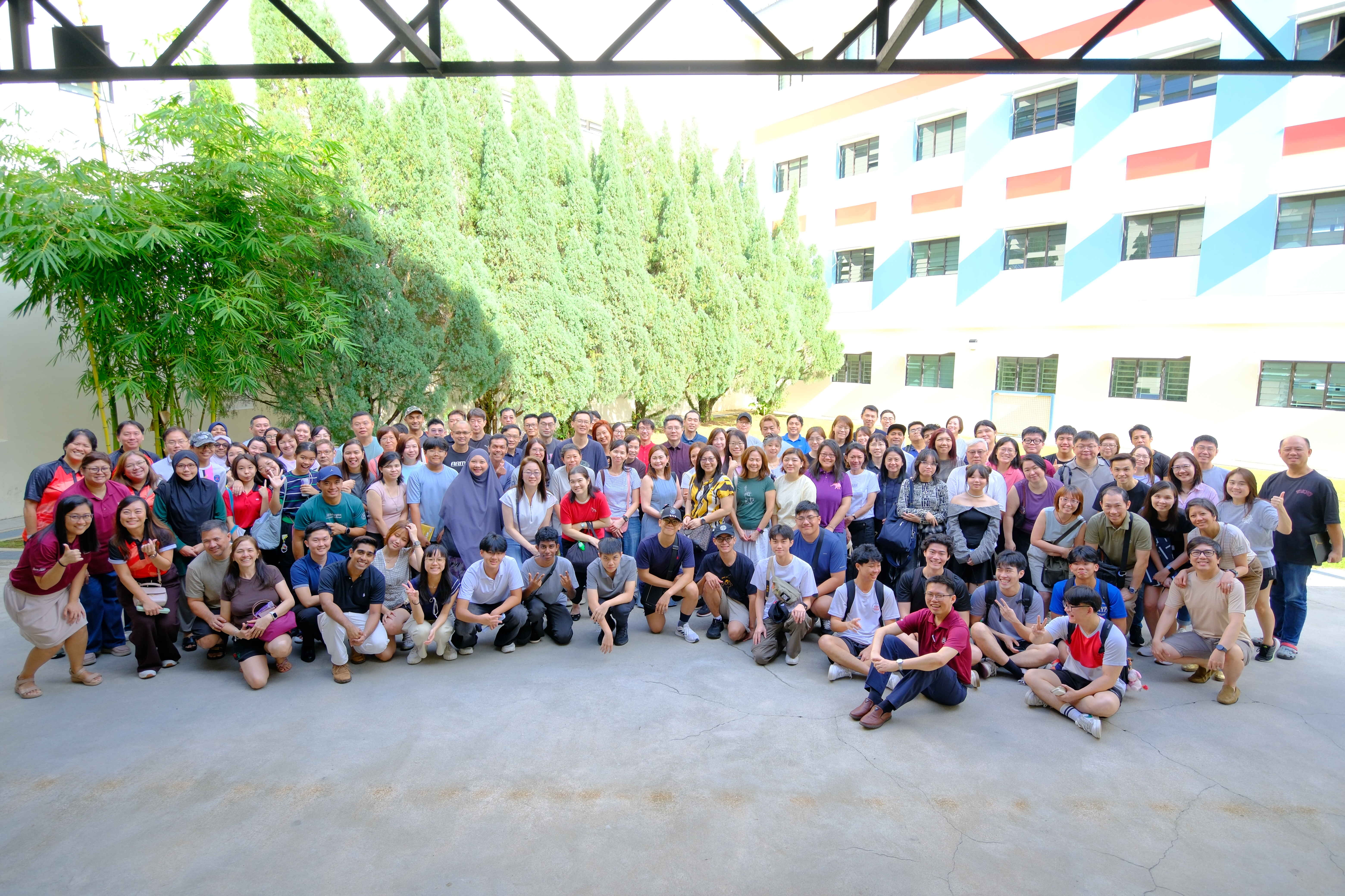 Large group of about 100 people posing for a photo outdoors near a colorful multi-story building and green trees.