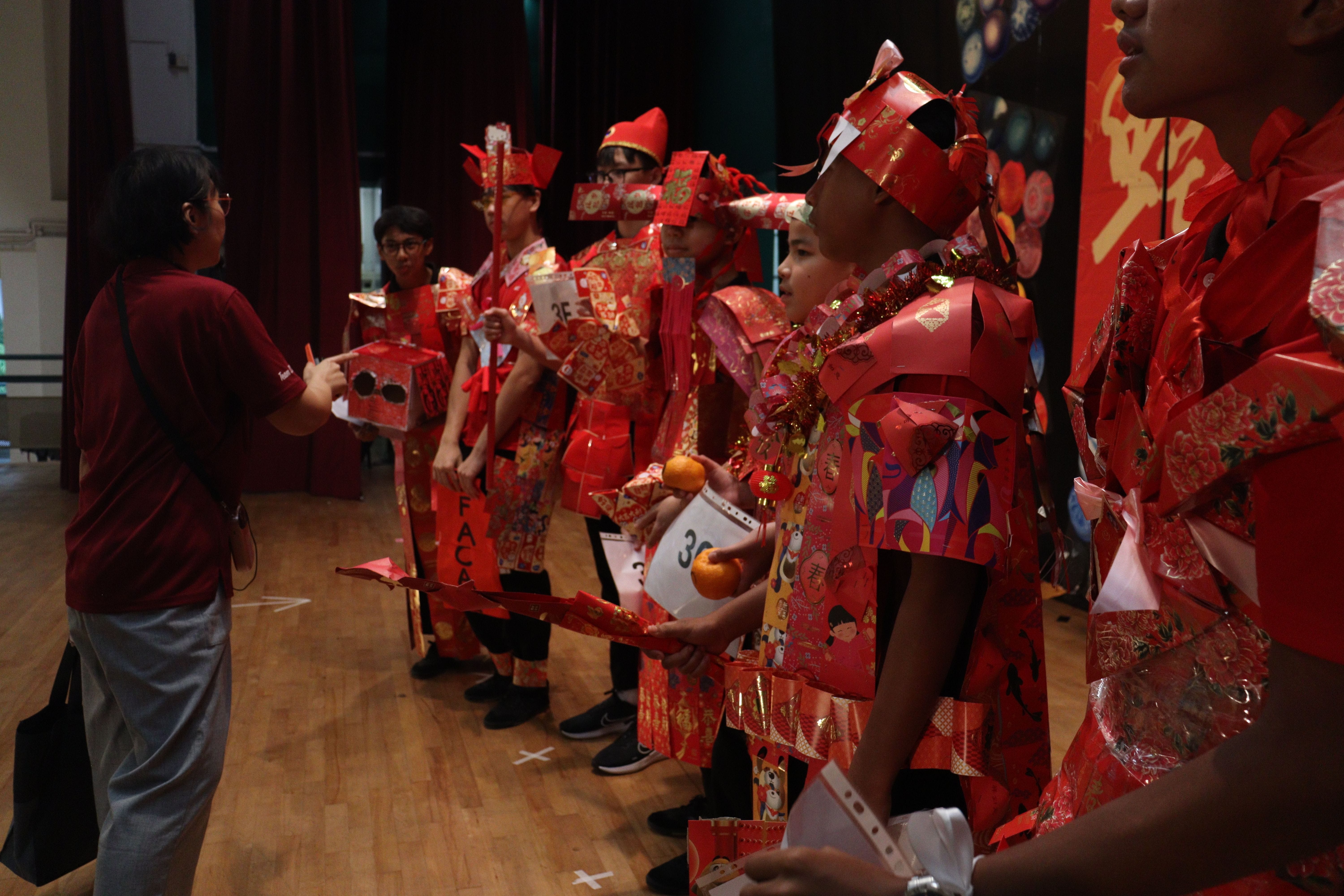Children wearing red paper costumes and hats stand in a line on a wooden stage during a Chinese New Year event.